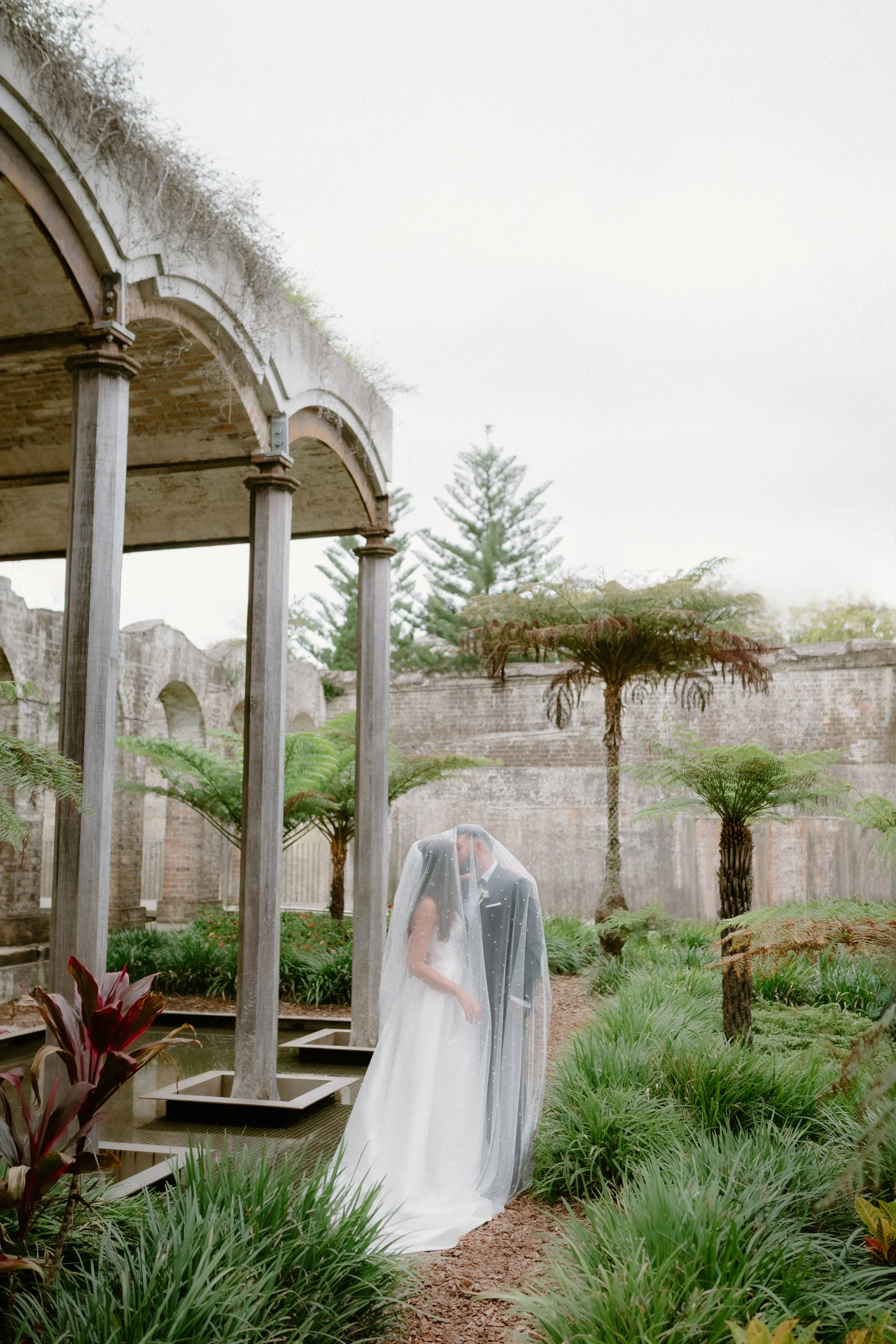 A bride and groom are standing close together in an outdoor garden setting, with the bride holding a bouquet and the groom gently holding her. They are covered with a sheer veil. The background features tall trees, stone walls, lush greenery, and an arched stone structure with columns.
