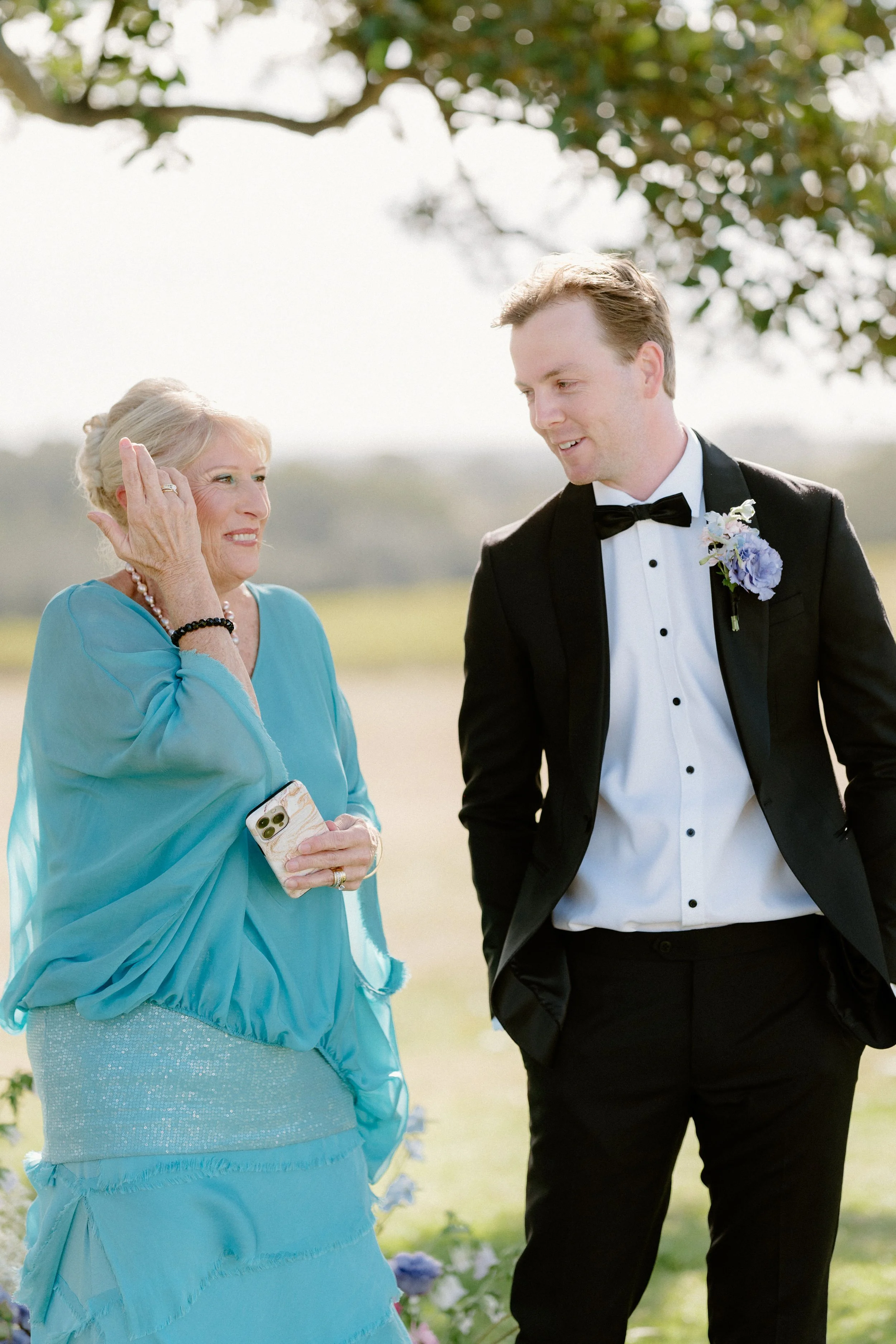 A woman in a light blue dress talking to a man in a black tuxedo outdoors during daytime. The woman is holding a phone and smiling, and the man is looking at her with a gentle smile. There are trees and greenery in the background.