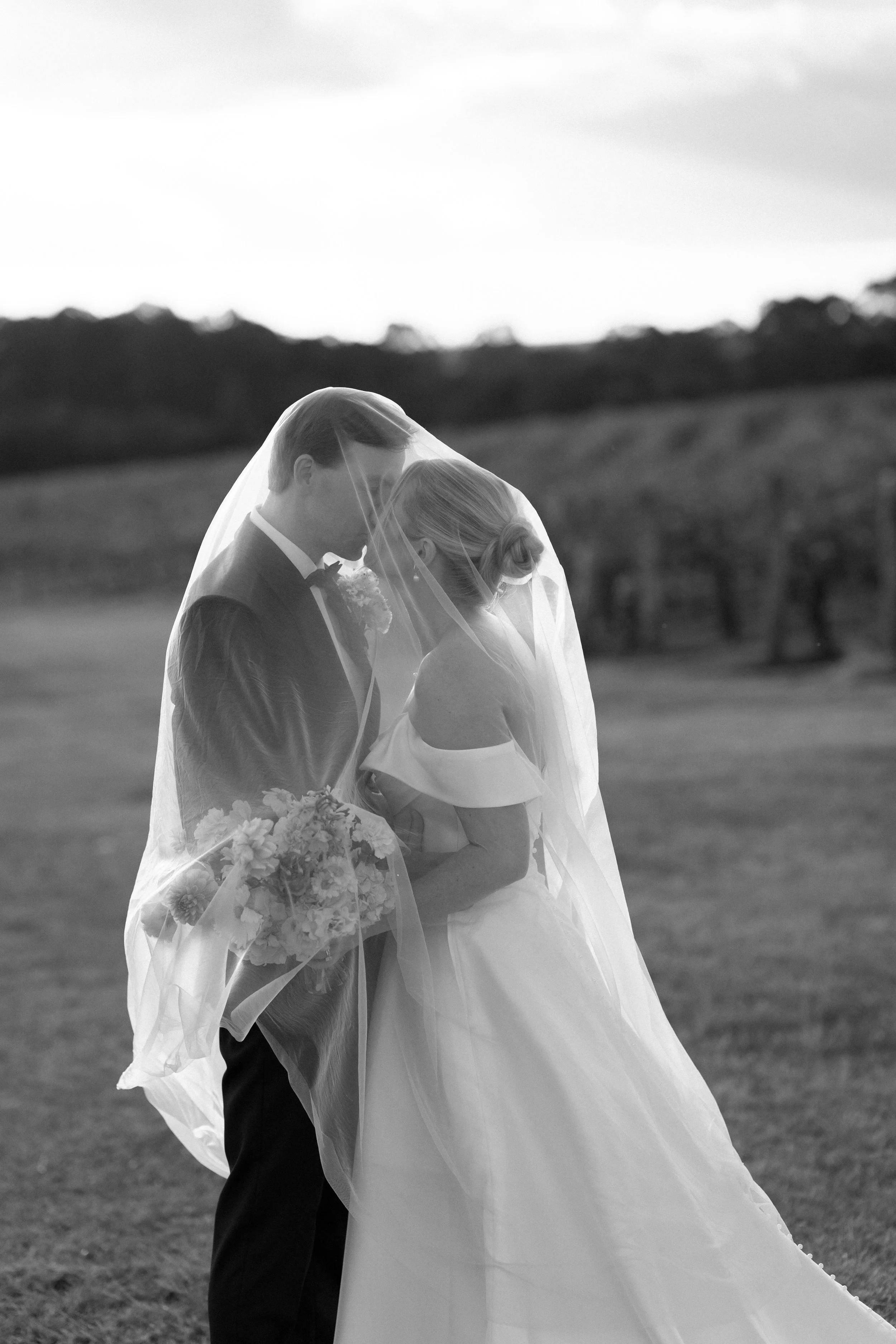 A black and white photograph of a bride and groom standing close in an outdoor setting, with the couple gazing at each other underneath the bride's veil. Bimbadgen Palmers Lane Wedding, Hunter Valley