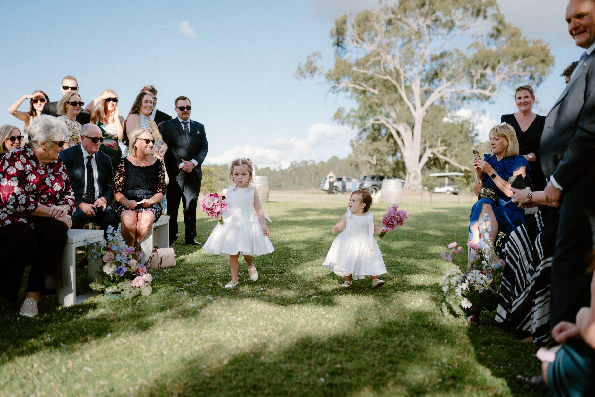 Children in white dresses holding pink bouquets walking down the aisle at an outdoor wedding ceremony with guests seated on both sides.