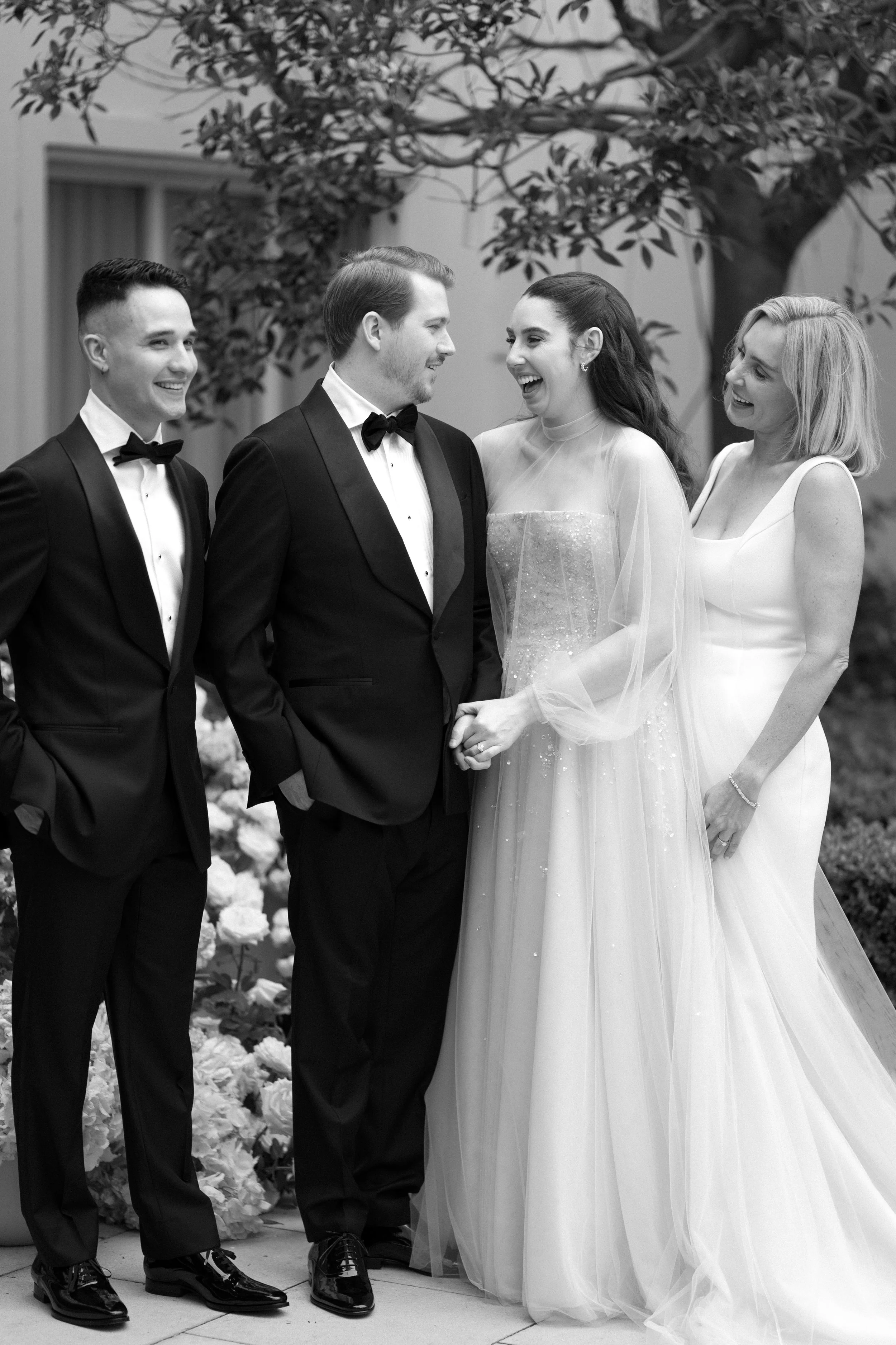 Black and white photo of a wedding party. Two men in tuxedos and bow ties, and two women in elegant dresses, standing outdoors with trees and flowers in the background. They are smiling and holding hands, celebrating a wedding.