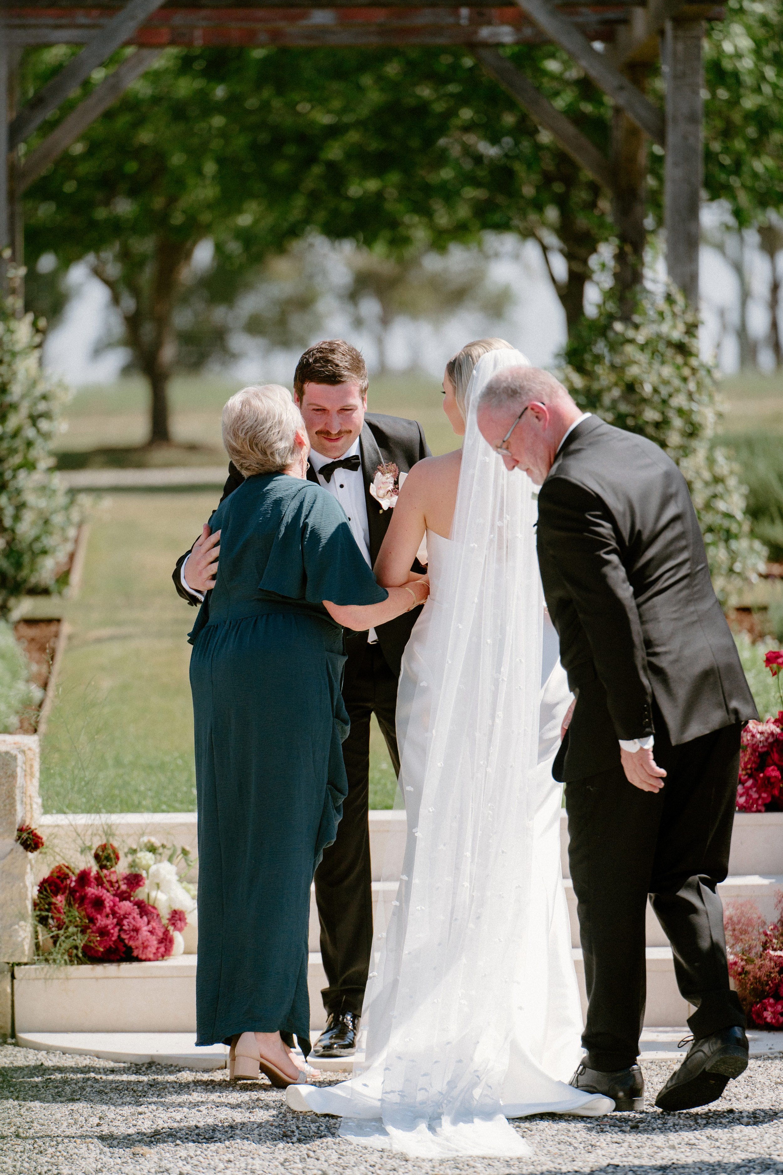 A bride and groom share a moment with two older adults during an outdoor wedding ceremony. The bride is in a white wedding dress with a veil, and the groom is in a tuxedo. The older woman is wearing a blue dress and the older man is in a black tuxedo
