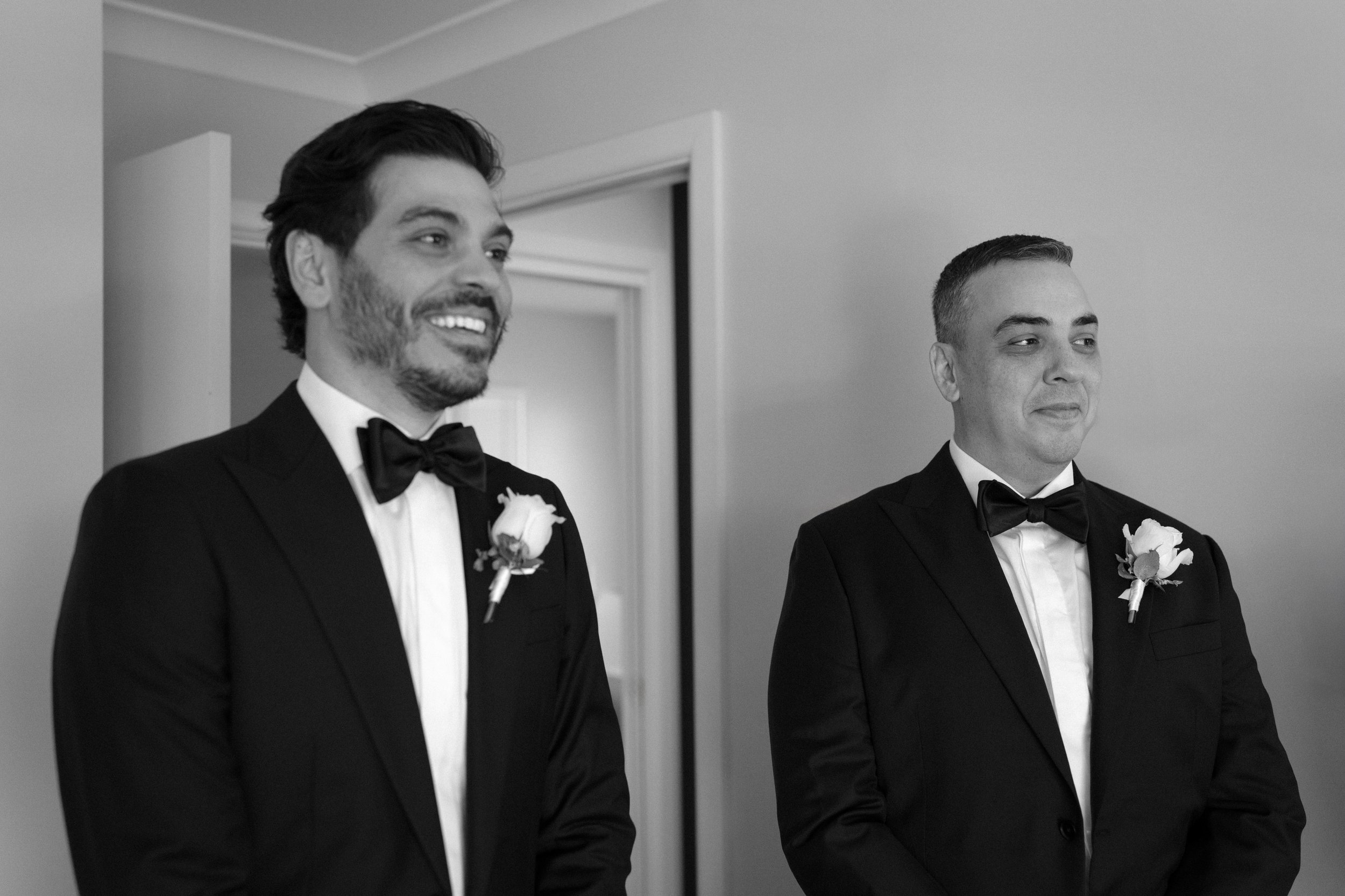 Black and white photo of two men in tuxedos with boutonnieres, standing indoors, one smiling and looking to the side, the other with a serious expression.