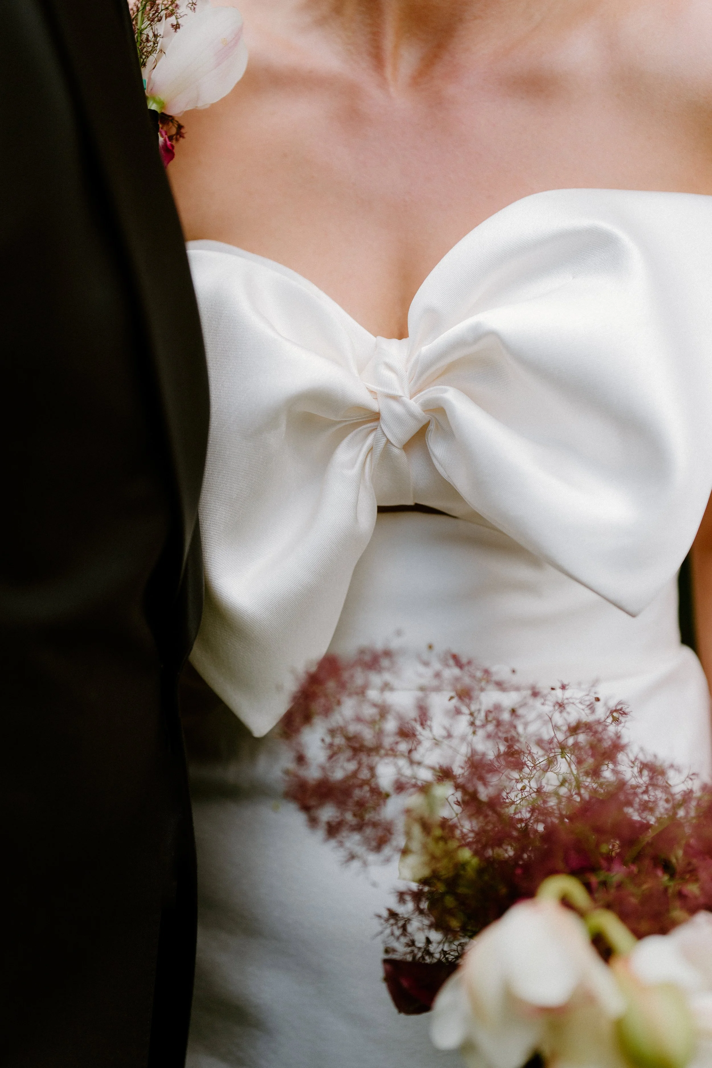 Close-up of a woman wearing a white dress with a large bow at the chest, holding a bouquet of flowers.
