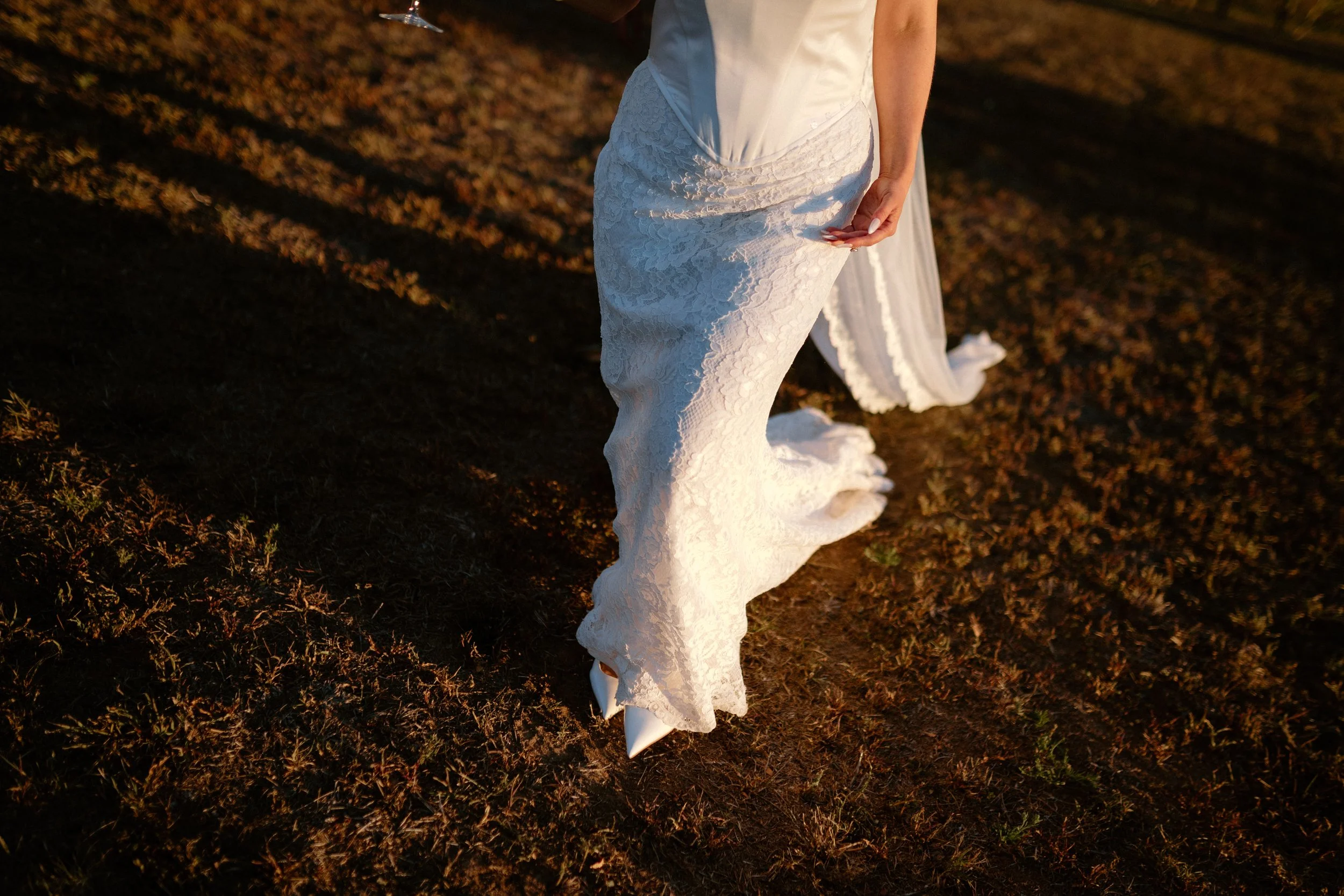 A person in a white lace dress standing outdoors on brown dirt ground, with the long train of the dress trailing behind them.