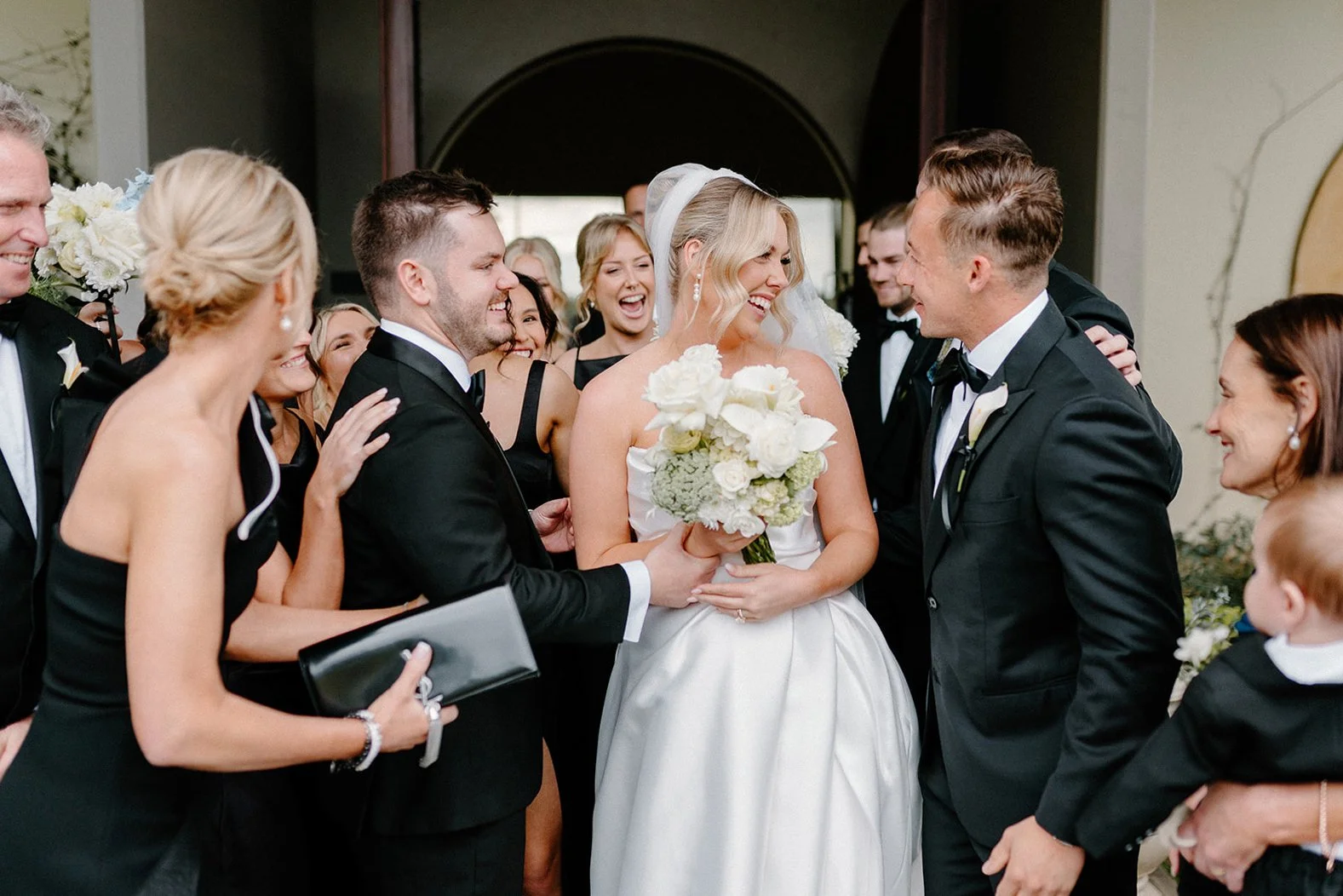 Bride and groom surrounded by wedding party, smiling and happy, during wedding celebration, woman holding bouquet of white flowers.