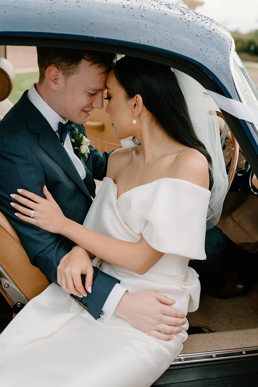 A newlywed couple sharing a close moment inside a vintage car, with the groom in a dark suit and the bride in an off-shoulder white dress, both smiling and touching foreheads.