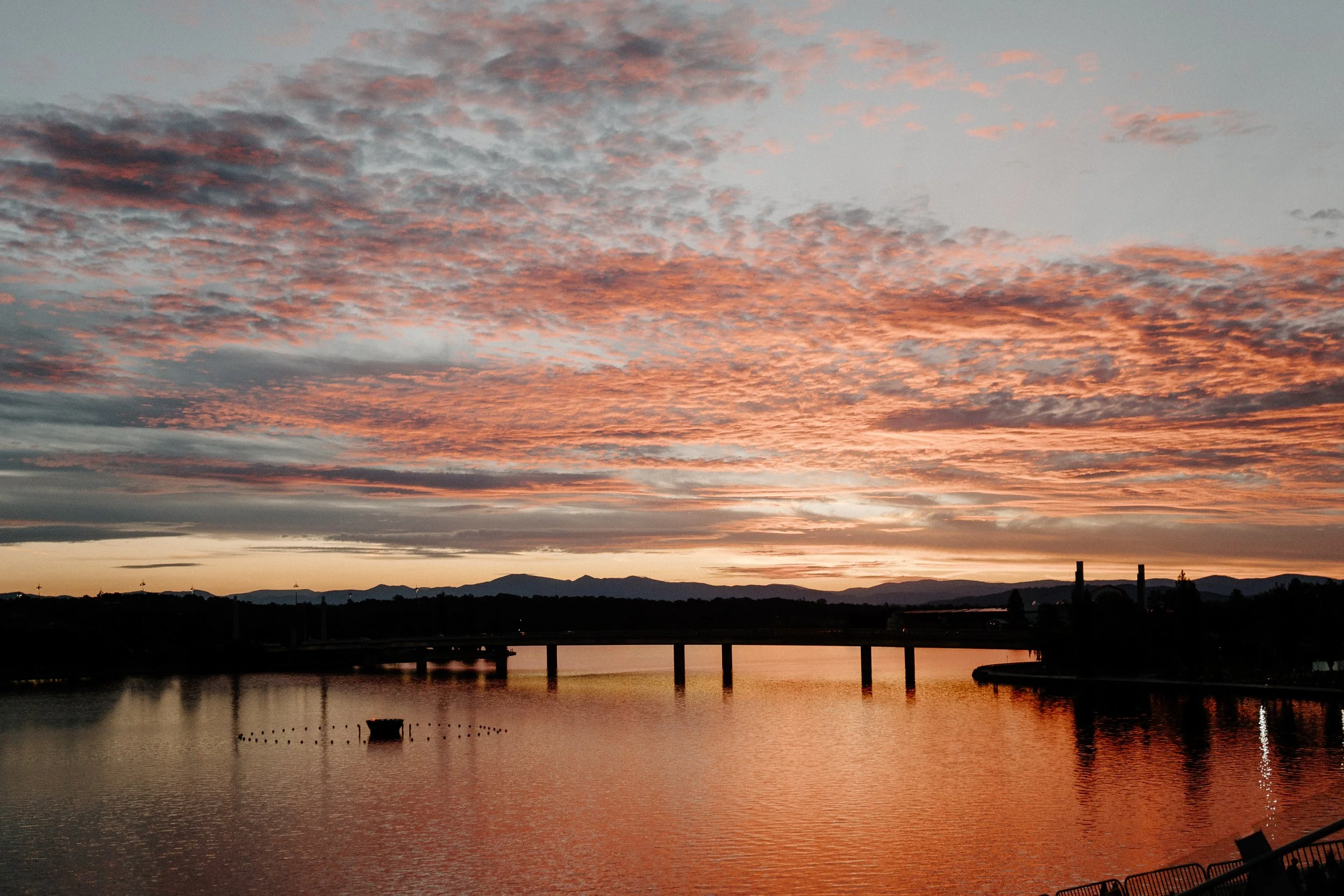 Sunset over a lake with pink and orange clouds, mountains in the background, and a bridge reflecting on the water.