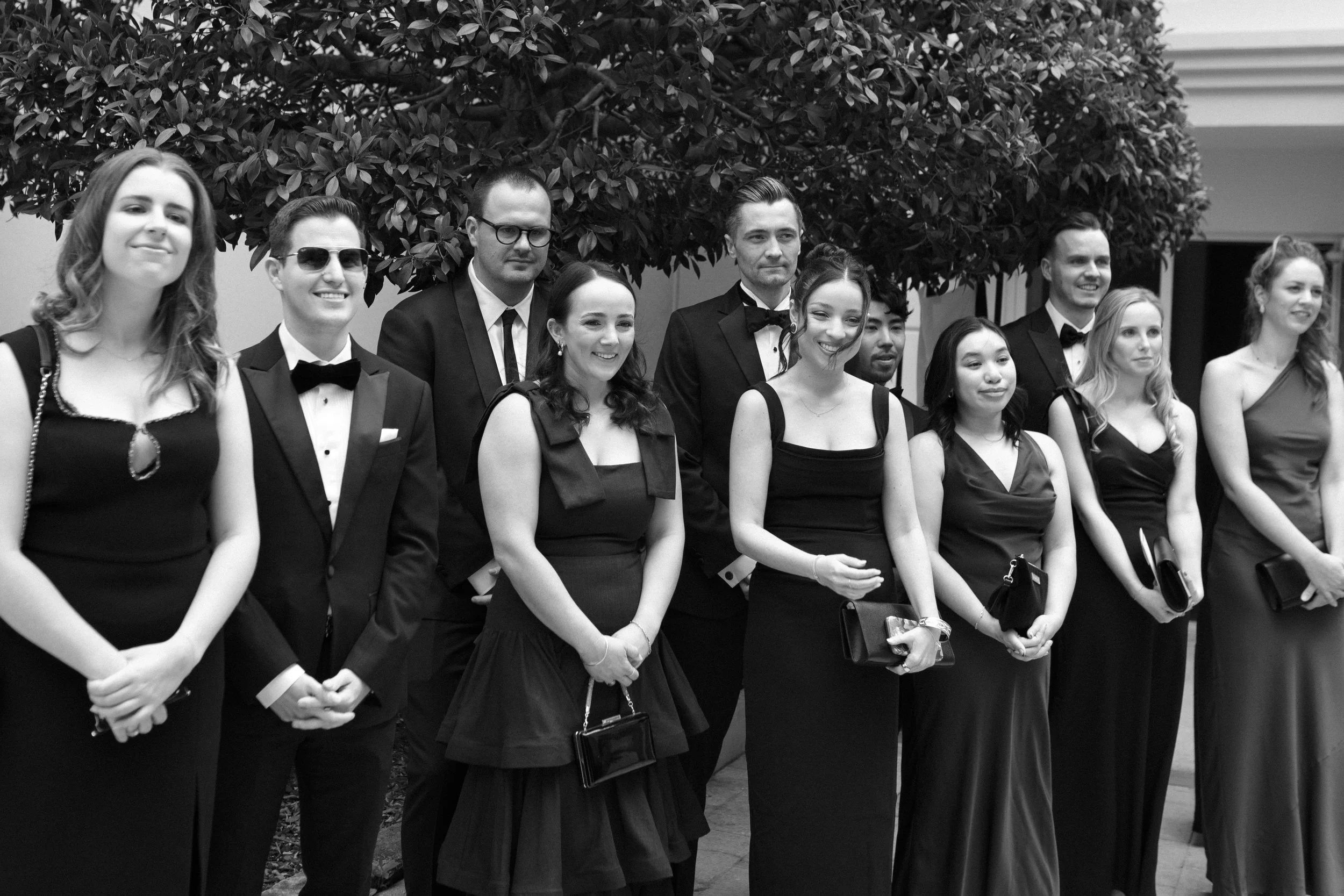 A group of people dressed in formal attire, standing outdoors in front of a large leafy tree, posing for a photo at a formal event.