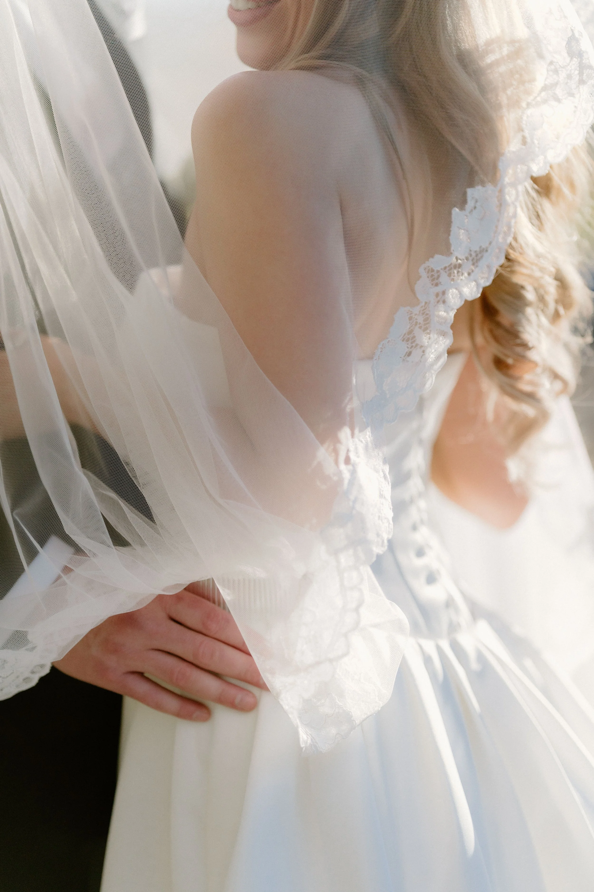Close-up of a bride in a white dress and lace veil with a hand on her waist, captured outdoors.