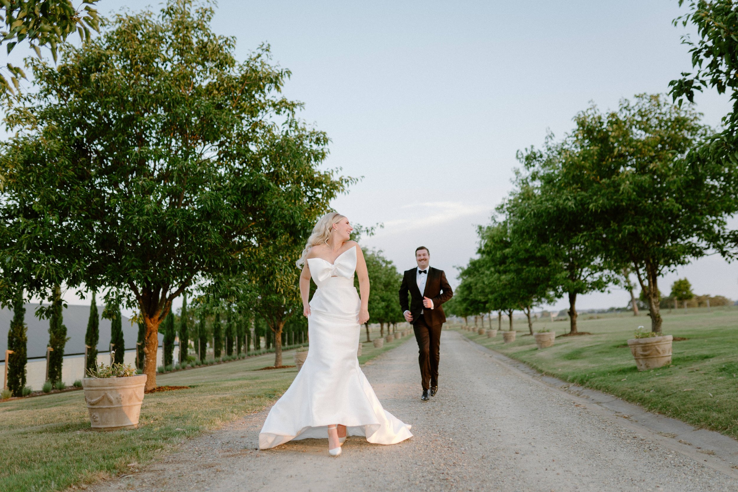 The Sienna, Hunter Valley Wedding Venue, Wedding Photos. A bride in a white wedding gown and a groom in a black tuxedo running along a tree-lined gravel path in a garden or park setting.