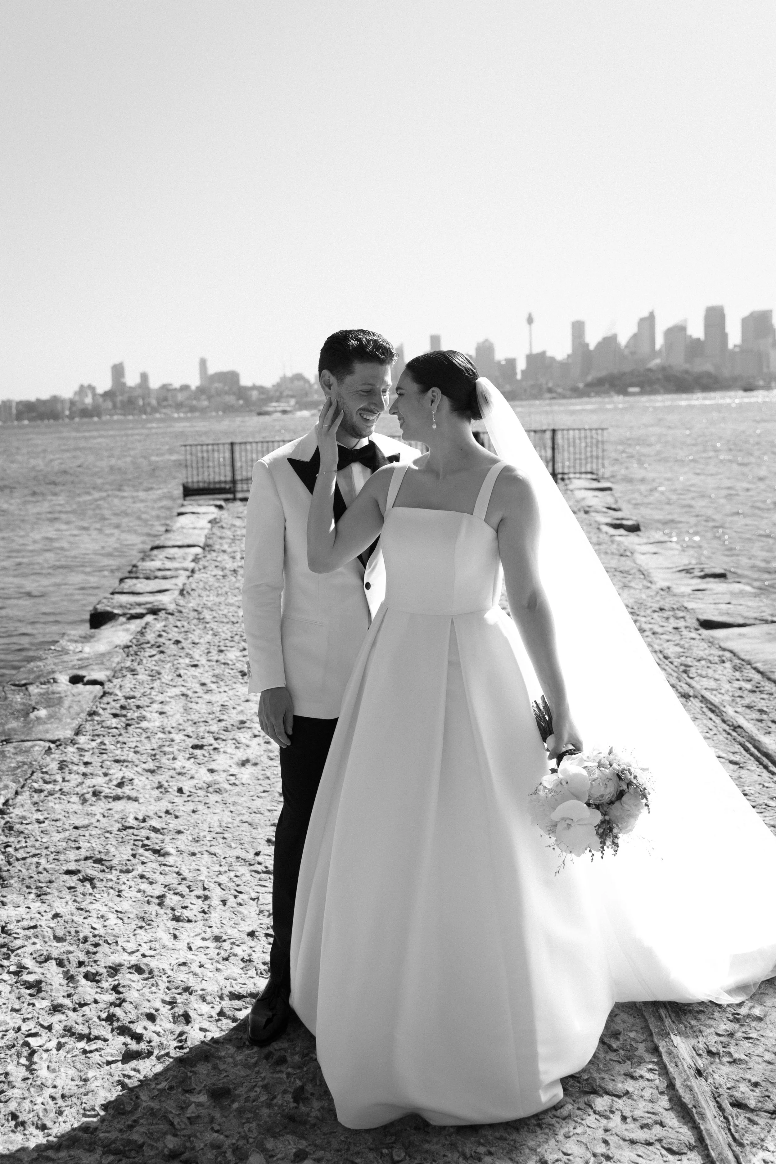 Sergeants Mess, Mosman Wedding Venue. Sergeants Mess Wedding Photos. Black and white photo of a bride and groom standing on a rocky shoreline with a city skyline in the background, smiling and touching foreheads.