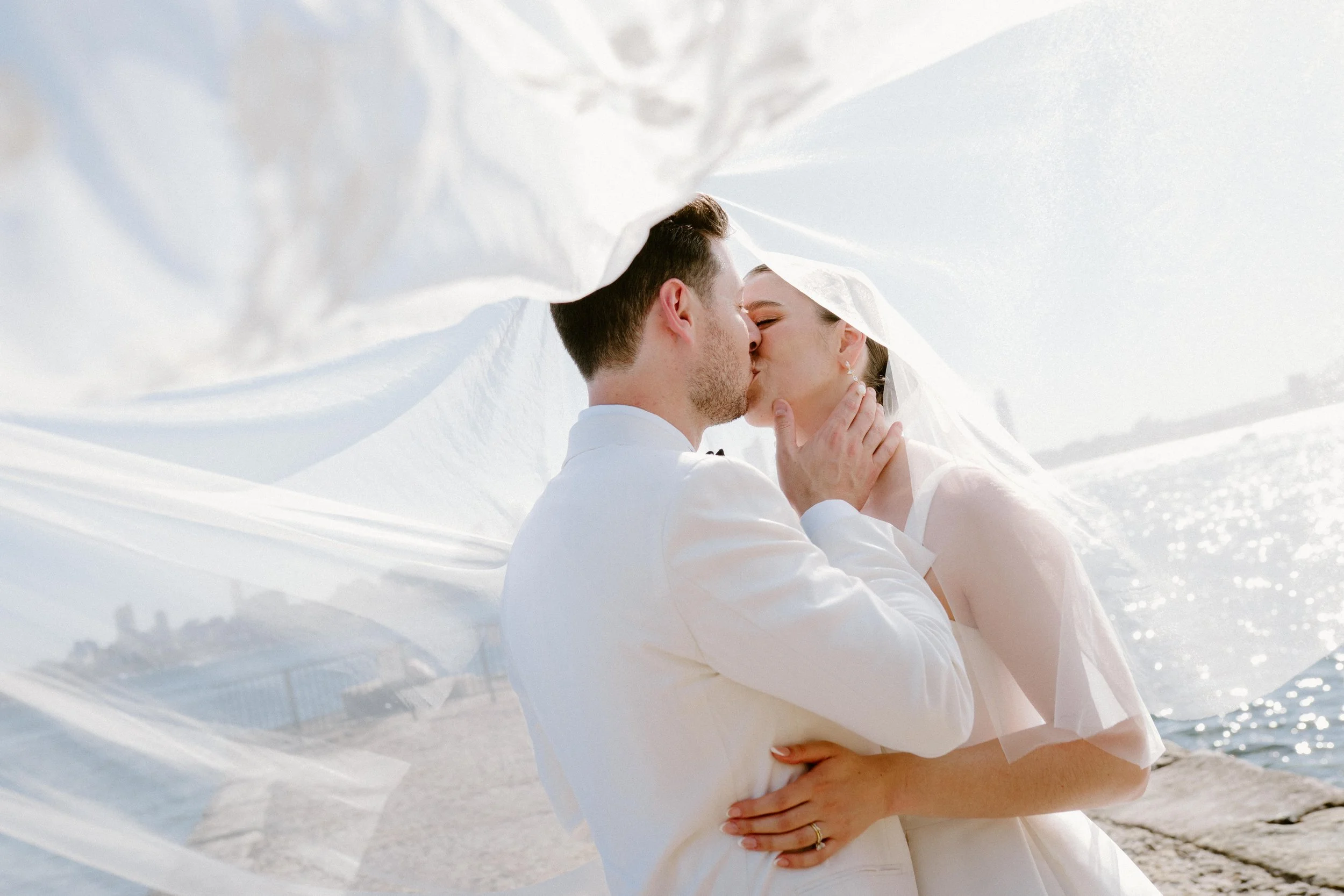A couple sharing a kiss outdoors near the water, with the bride's veil flowing around them.