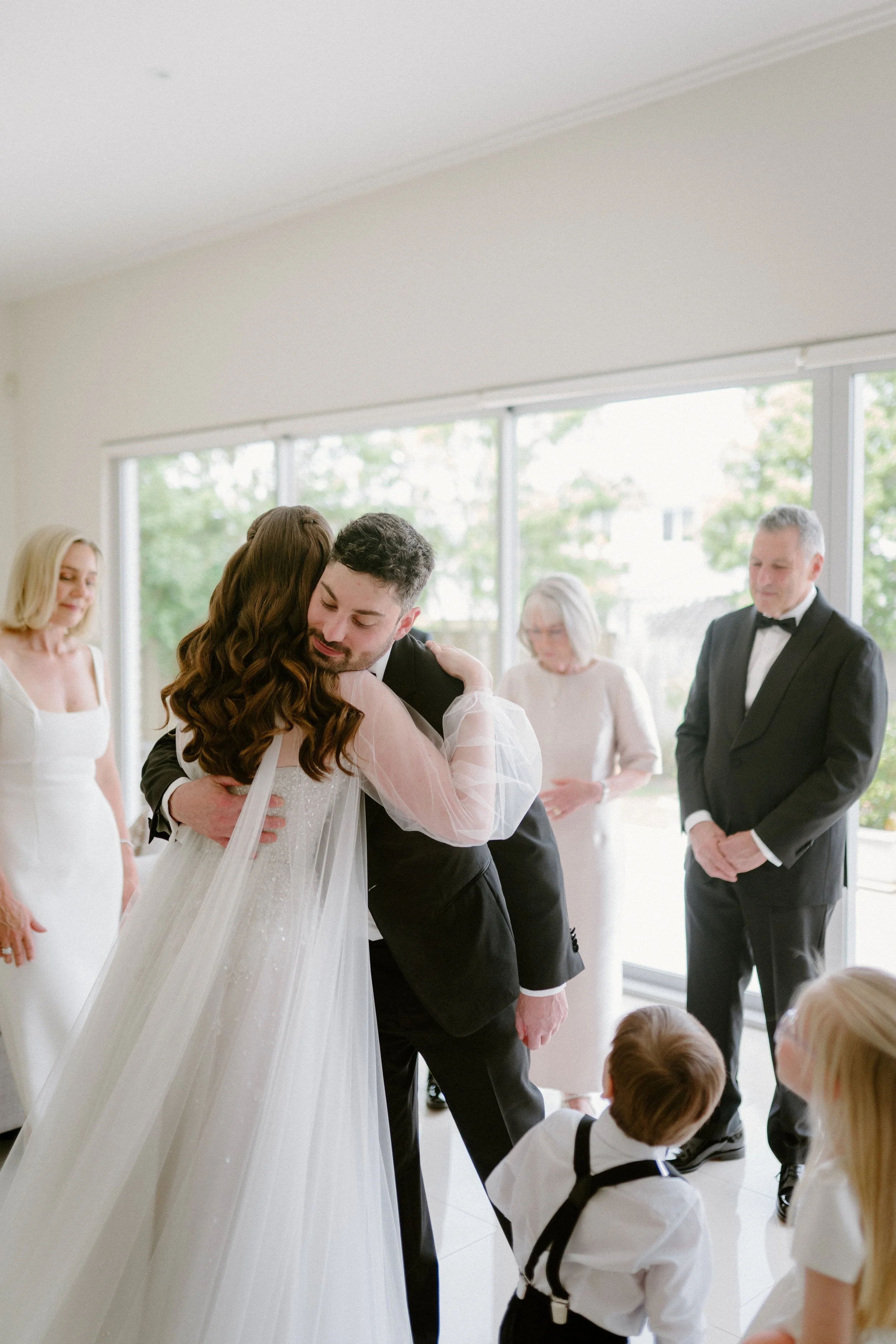 A bride and groom share an embrace during their wedding ceremony, with guests looking on in a bright indoor setting with large windows and greenery outside.