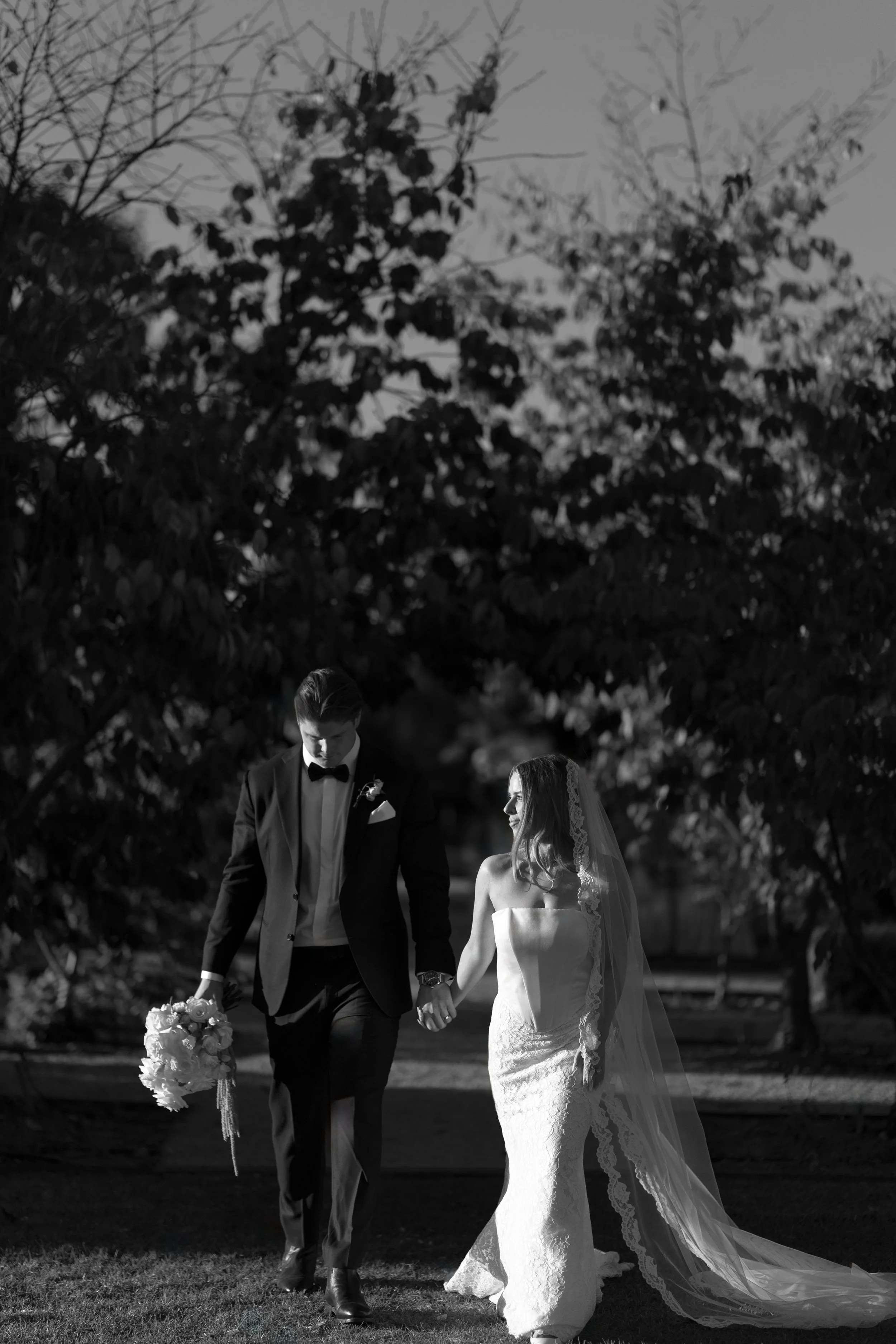 A black and white photo of a bride and groom walking hand in hand outdoors. The groom is wearing a tuxedo and the bride is in a wedding gown with a train and veil. They are surrounded by trees.