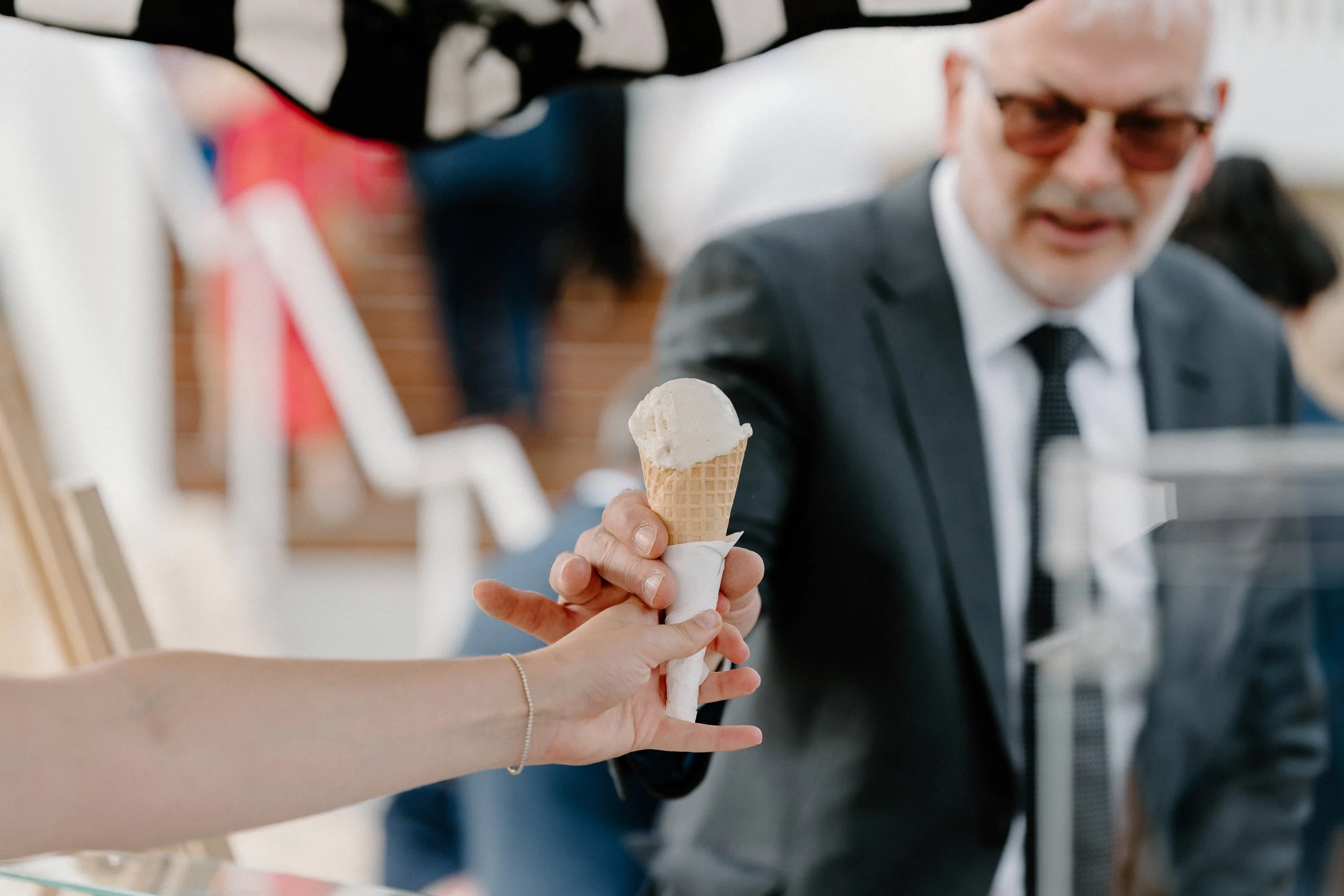 Person in a black suit and glasses receiving a scoop of vanilla ice cream in a cone from another person with a bracelet on their wrist, in an indoor setting.