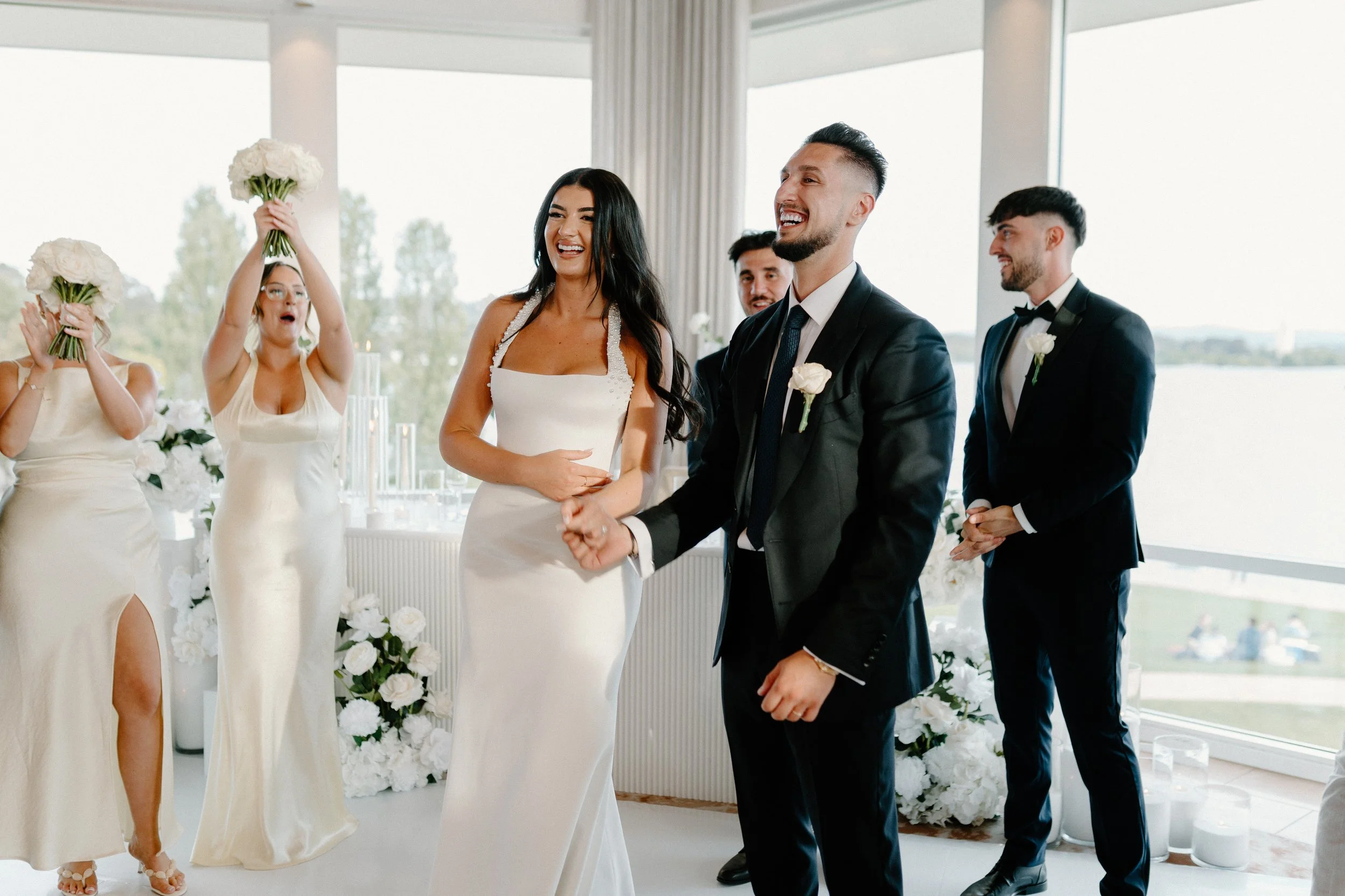 A wedding ceremony with a bride and groom smiling and holding hands, surrounded by bridesmaids and groomsmen dressed in formal attire, with a bright and airy backdrop of large windows and outdoor scenery.