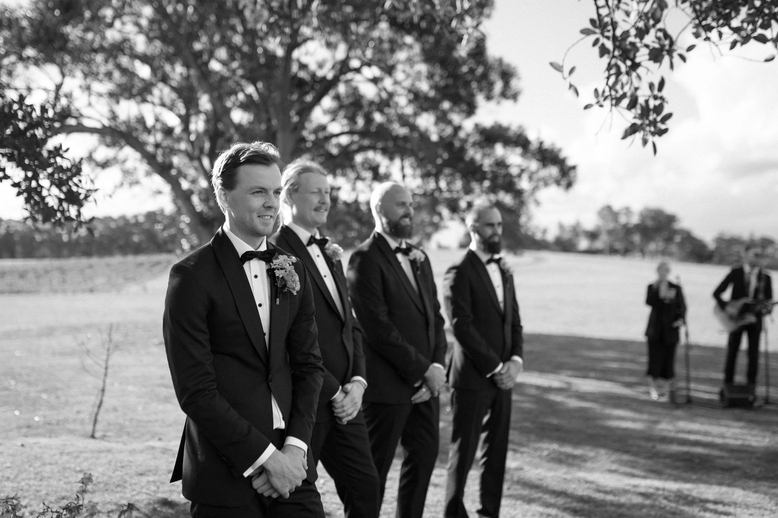 A group of men in tuxedos standing outdoors at an event, with trees and a grassy area in the background.