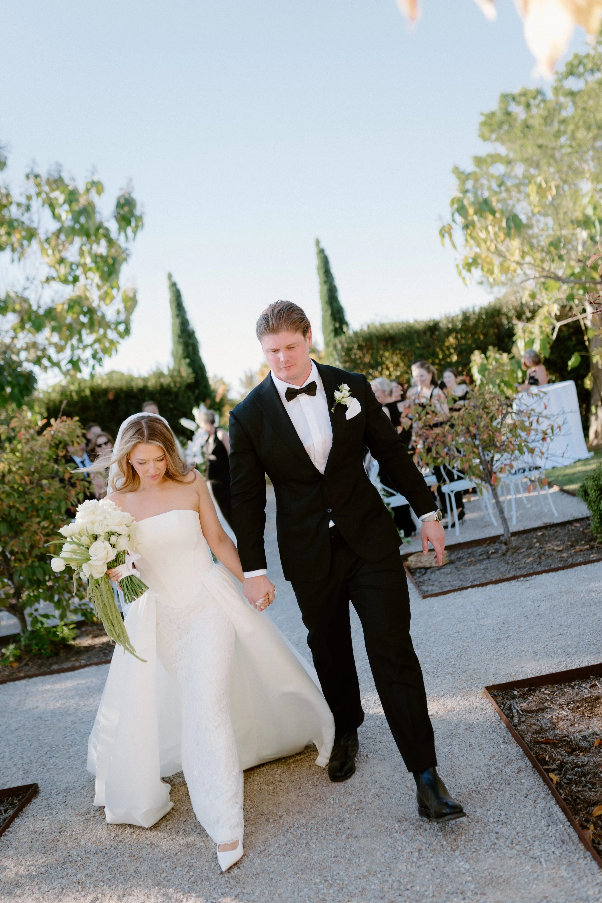 A bride and groom walking hand in hand outdoors during a wedding celebration, with guests and greenery in the background.