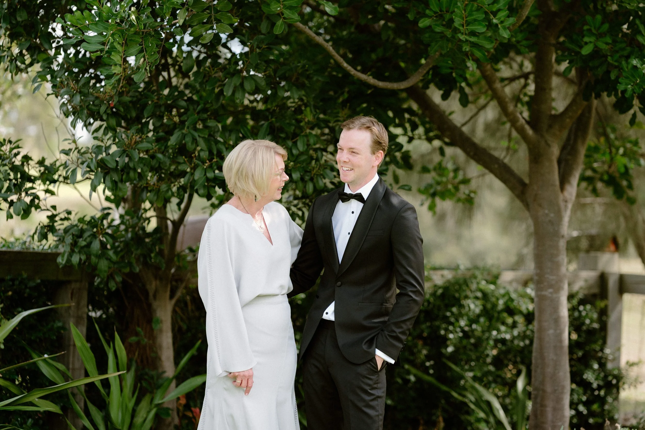 A woman and a man sharing a smile outdoors, dressed formally, standing near trees and greenery.