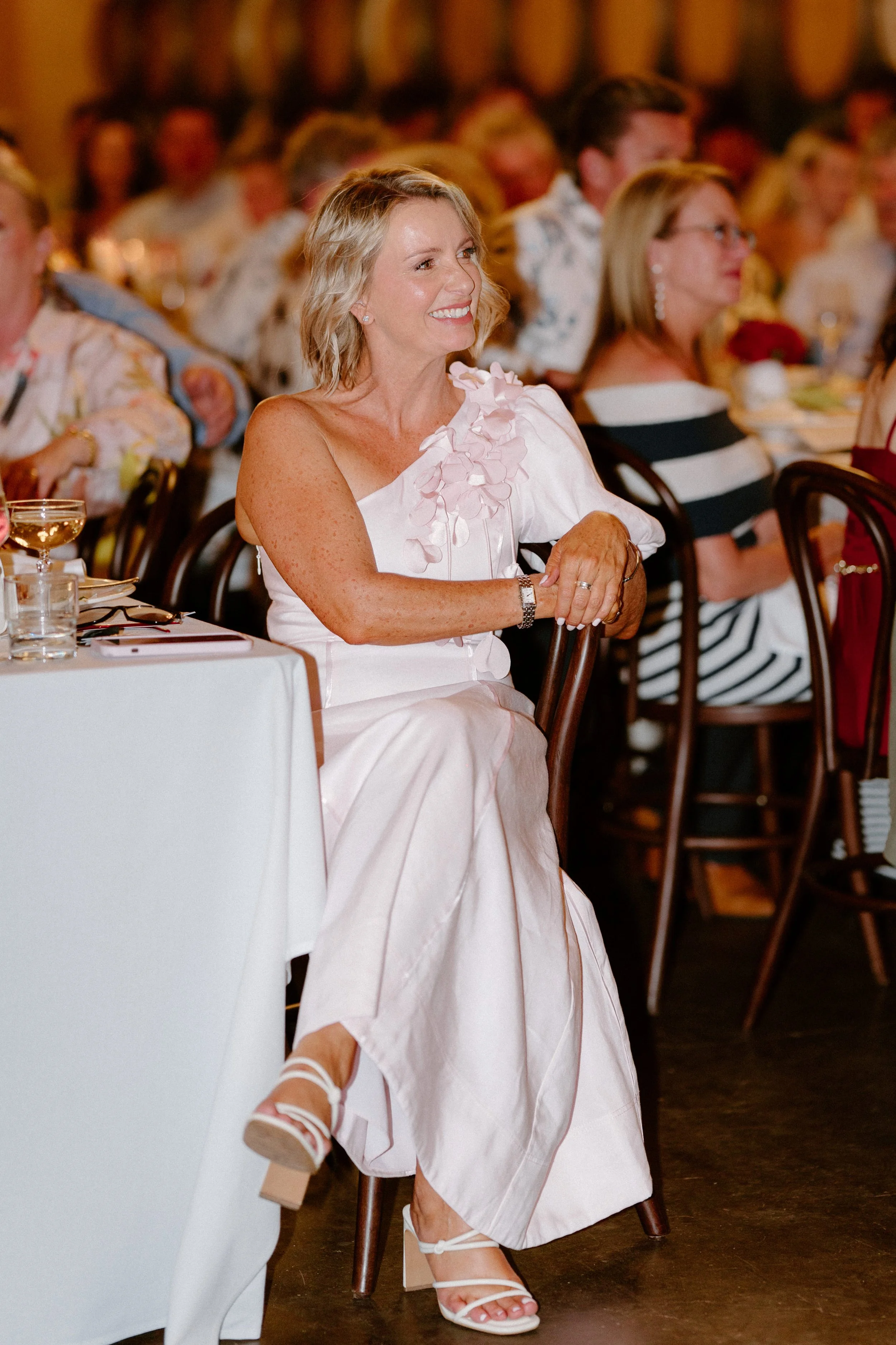 A woman with blonde hair, wearing a white dress with floral details, is sitting at a table during an event or celebration, smiling and looking to her left.