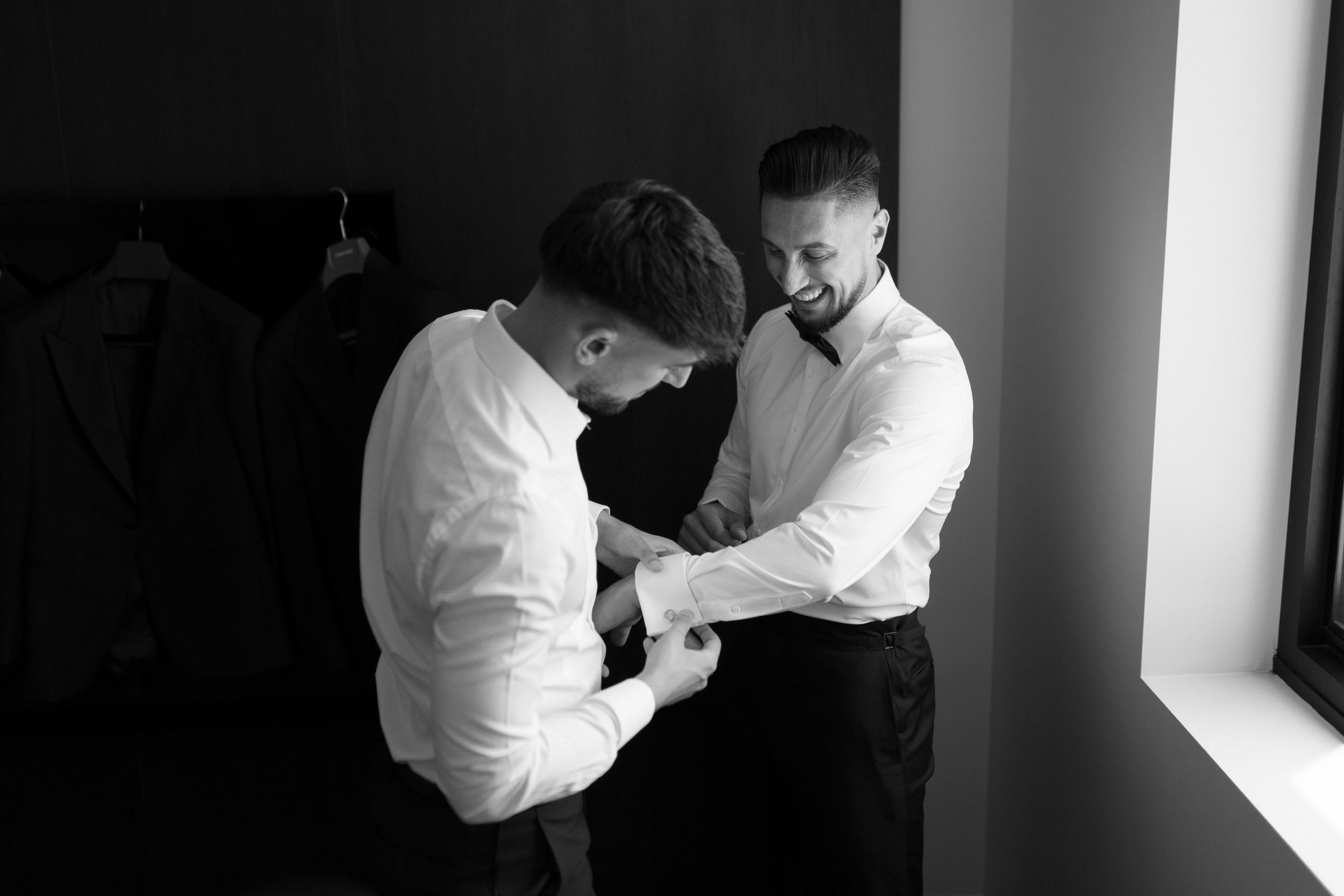 Two men dressed in formal white shirts, one adjusting his cufflinks while the other smiles and watches, in a room with dark walls and a window.