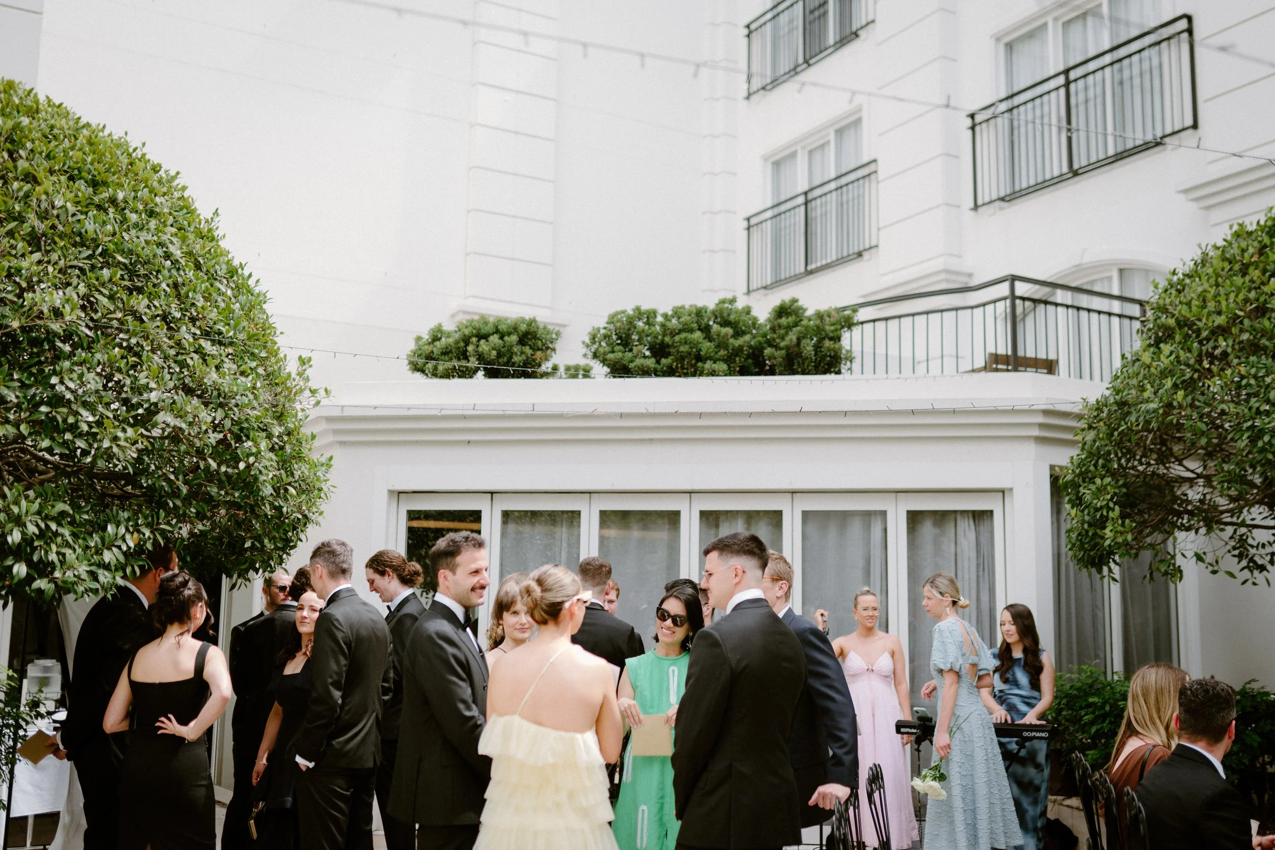People in formal attire gathered outdoors for a wedding or event, with women in dresses and men in suits, on a patio surrounded by greenery and white building.