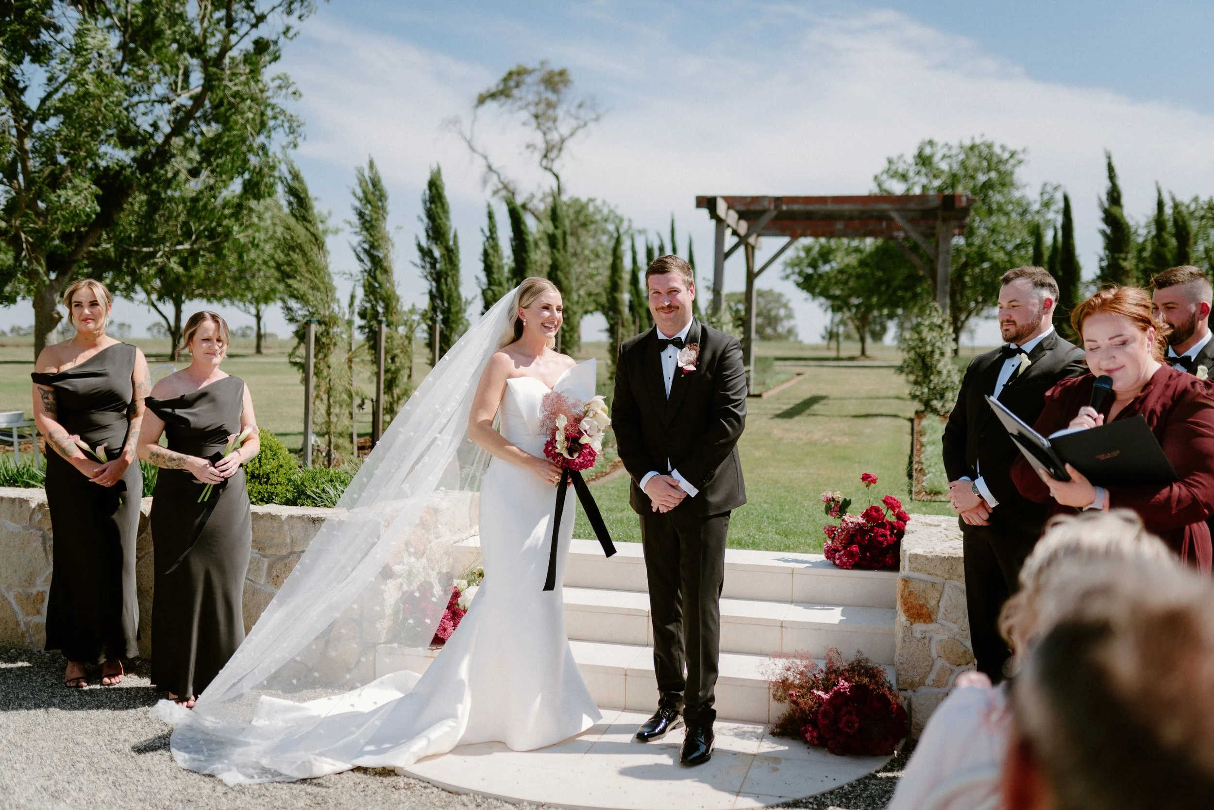 A bride and groom exchanging vows outdoors at a wedding ceremony surrounded by bridesmaids and groomsmen, with blue sky and green trees in the background.