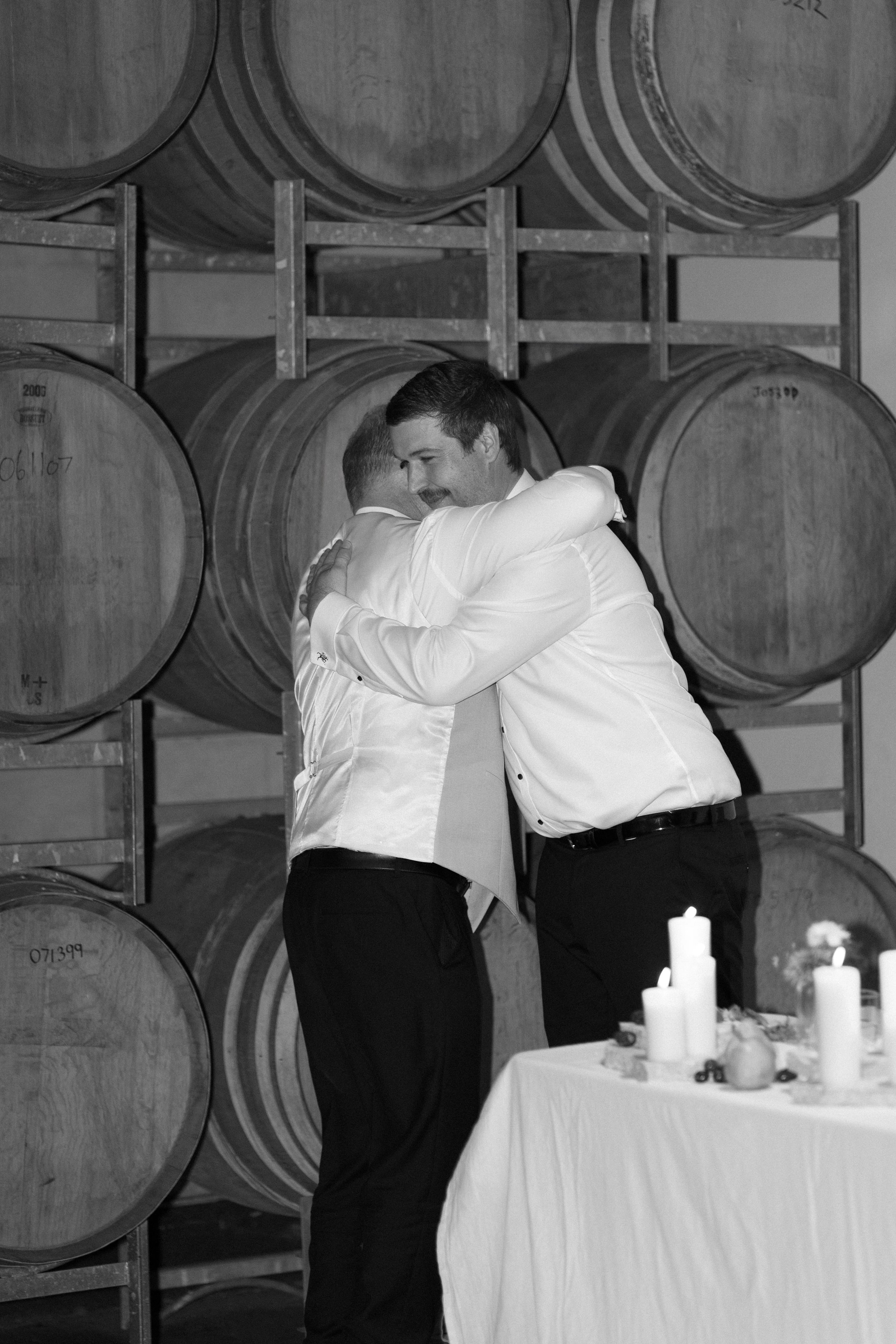 Two men hugging at a wedding reception in front of wine barrels, with a table of candles and decorations nearby.