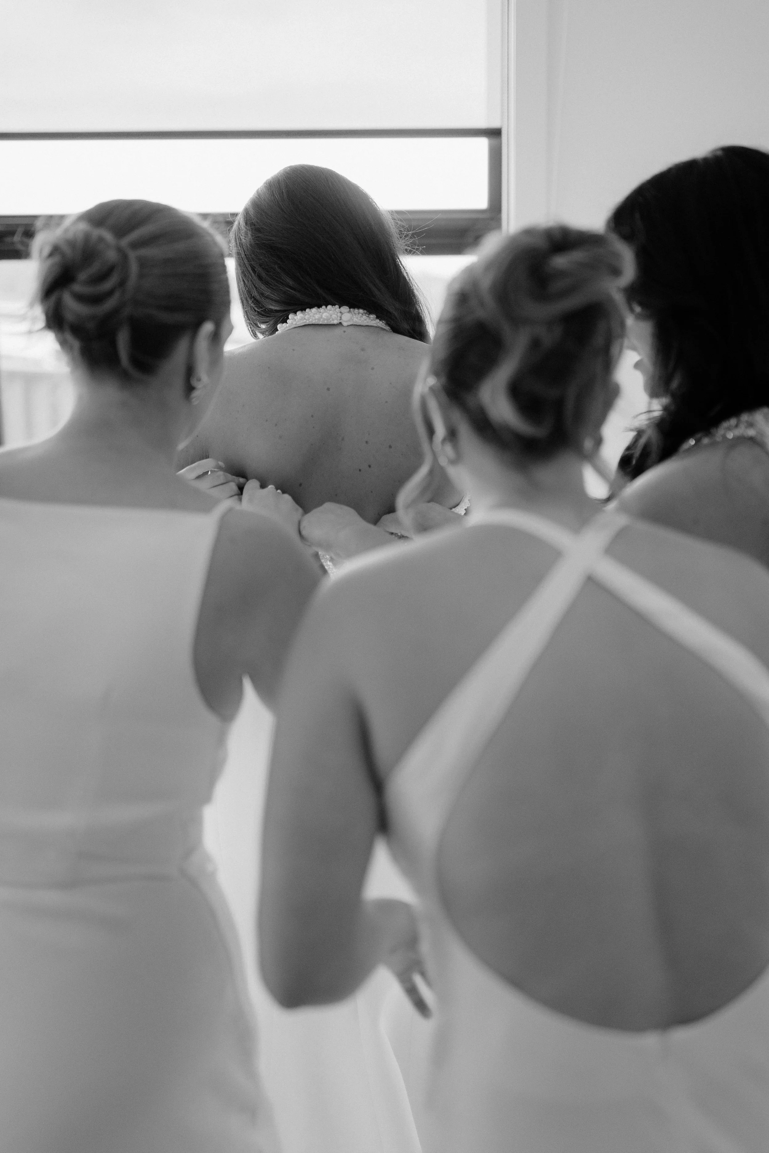 Black and white photo of women in elegant dresses, seen from behind, sitting by a window, with one woman having her back turned, surrounded by women helping her with a dress or accessories.