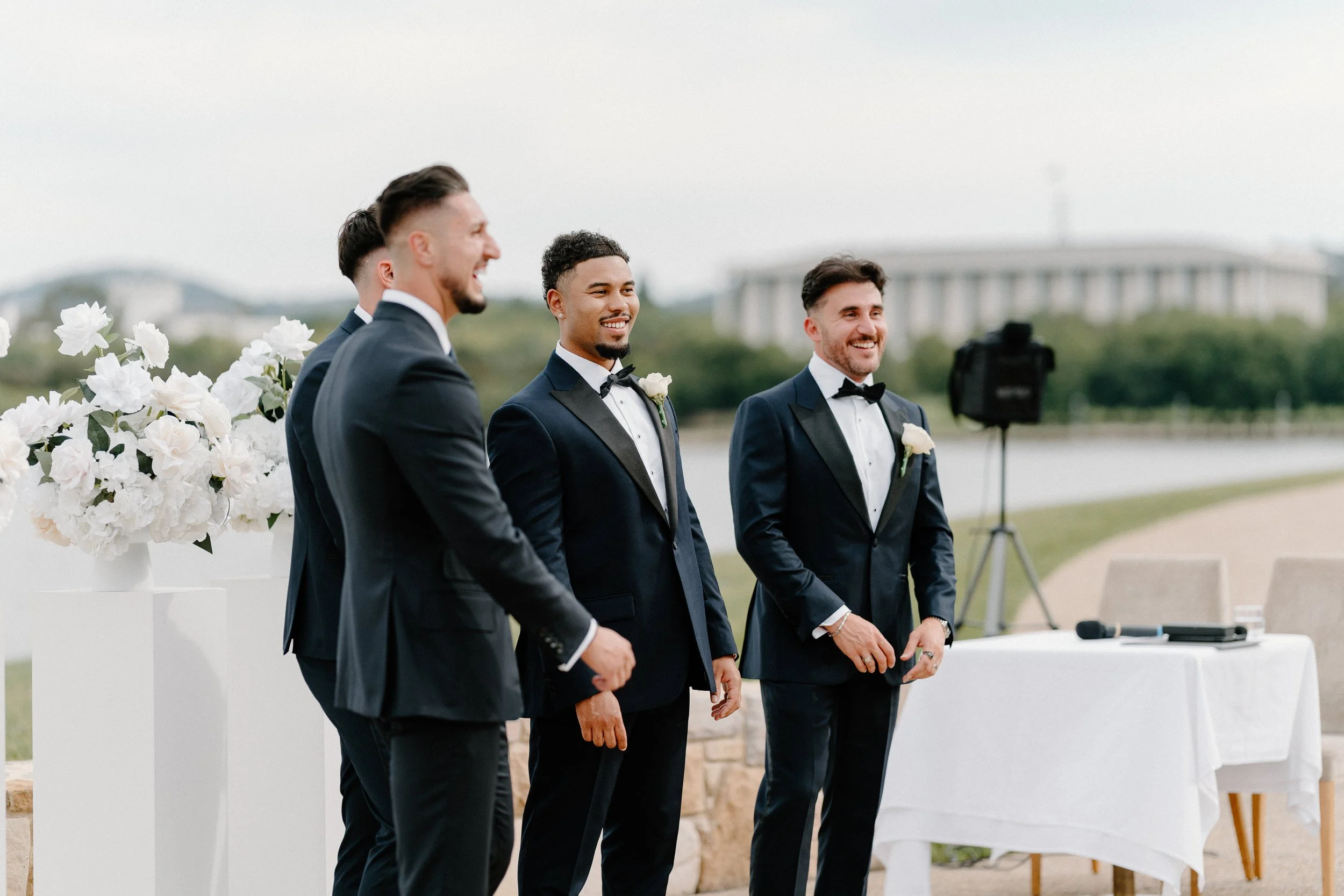 Four men in tuxedos at a wedding ceremony outdoors, smiling and standing next to a table with flowers, with a body of water and a building in the background.