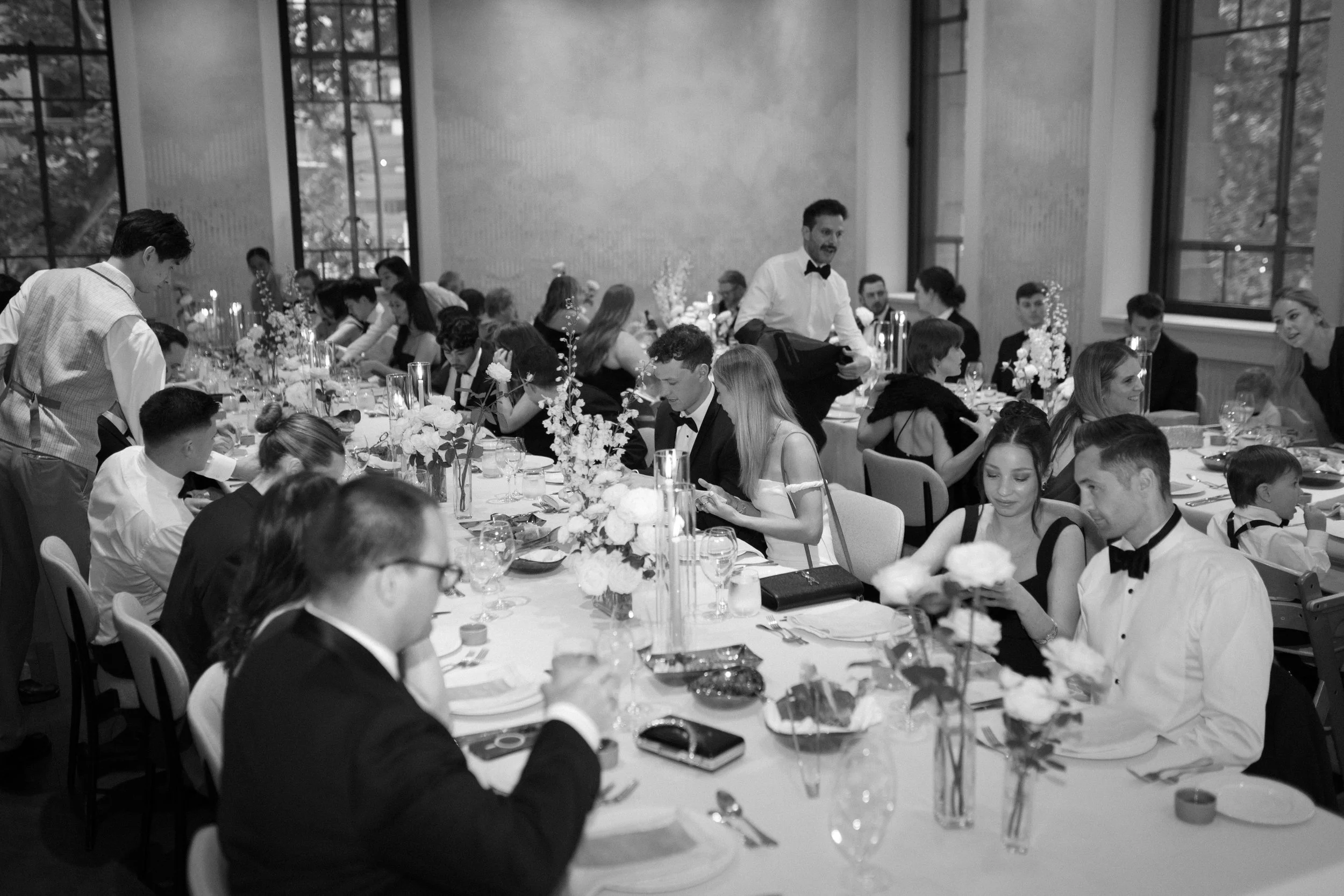 Black and white photo of a formal banquet with guests seated at a long table decorated with flowers and candles, with waiters serving. Large windows are visible in the background.