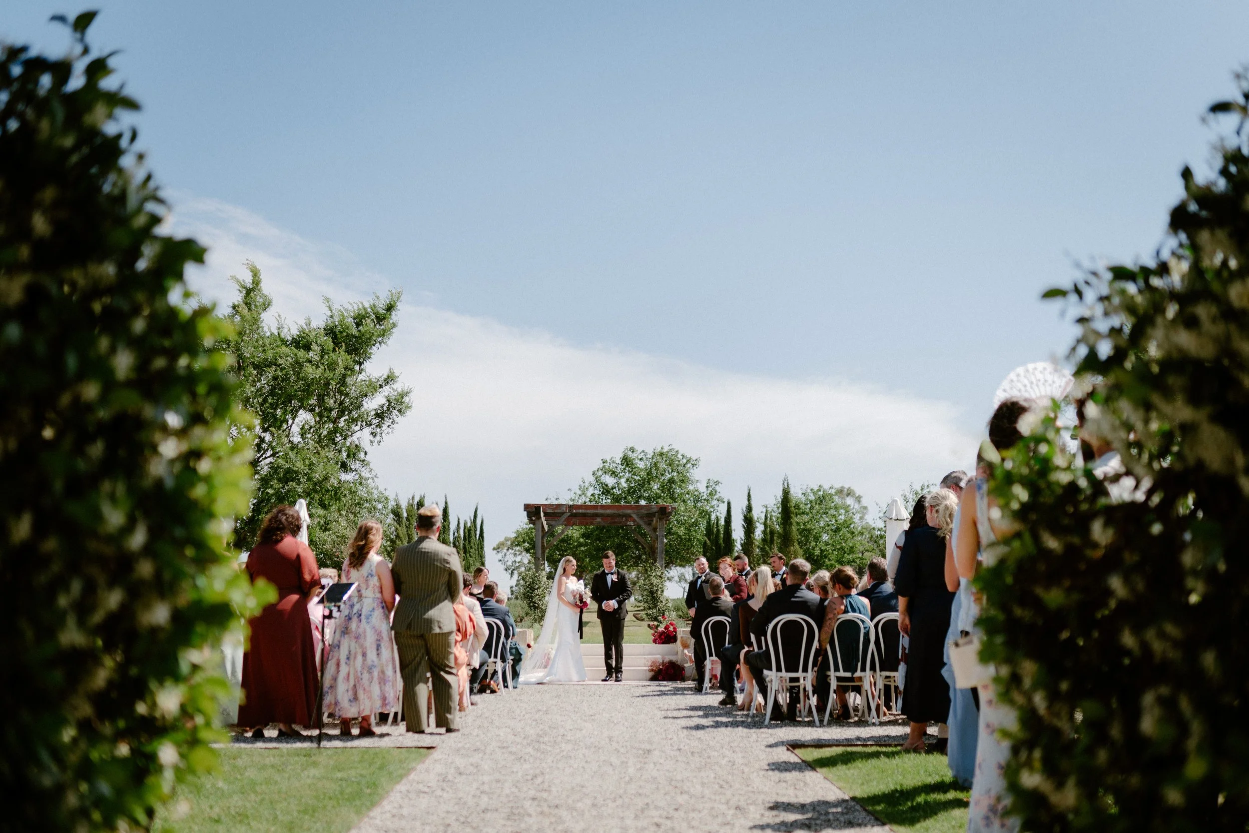Bride and groom standing under a wooden arch during outdoor wedding ceremony, guests seated on either side, bright sunny day with blue sky and green trees in background.