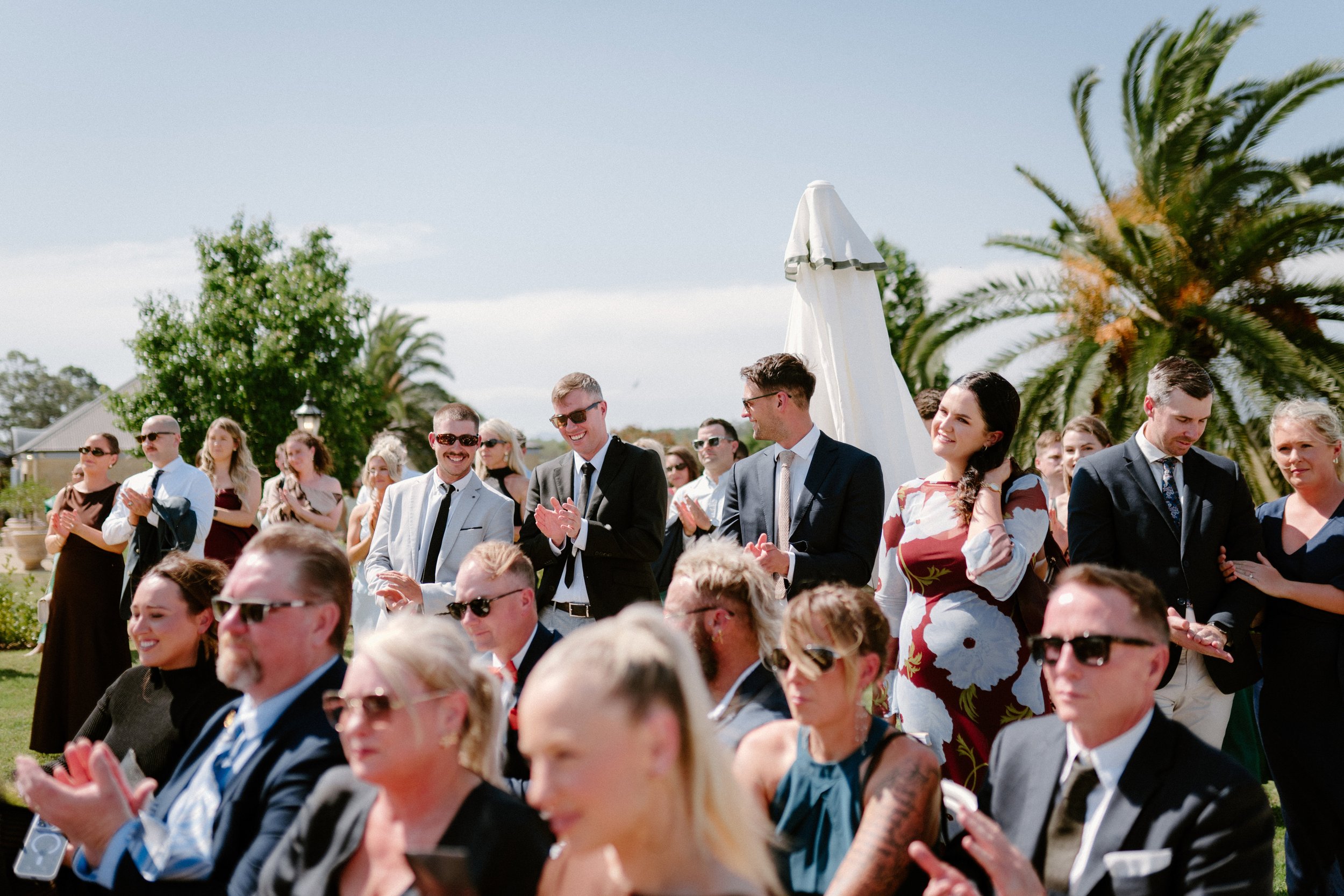 People attending an outdoor event, dressed in formal attire, clapping and smiling.