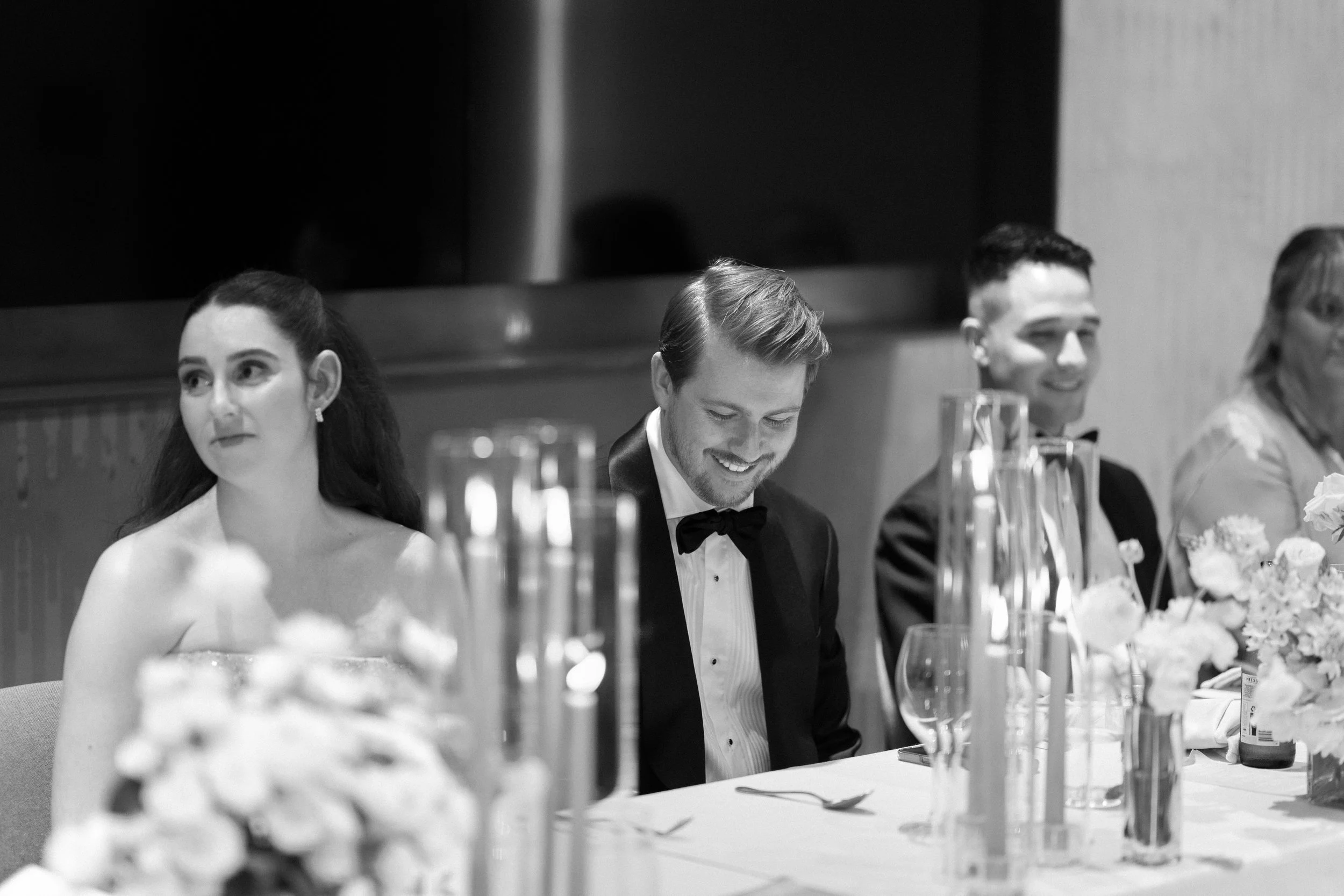 People sitting at a formal event, dressed in tuxedos and evening gowns, with flower arrangements and table settings in front of them.