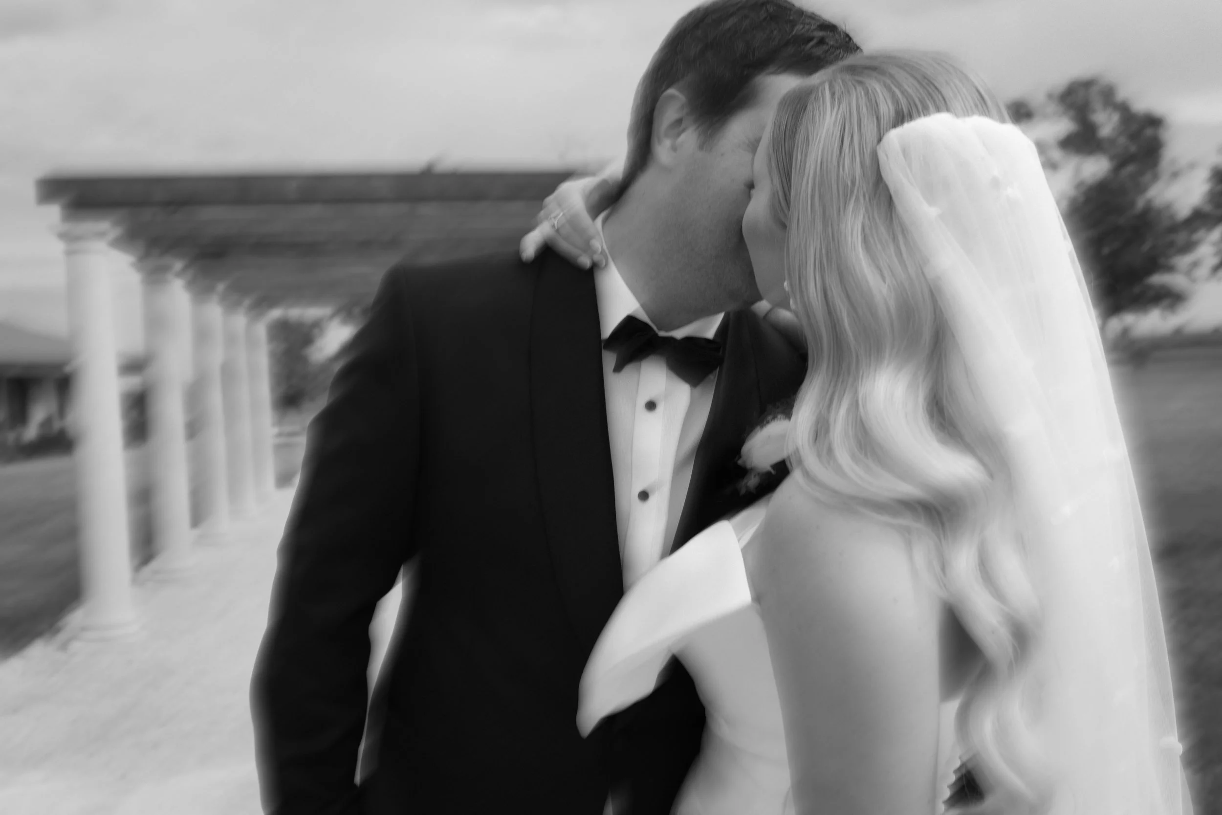 The Sienna, Hunter Valley Wedding Venue, Wedding Photos. A black and white photo of a bride and groom sharing a kiss outdoors during their wedding, with classical columns and trees in the background.