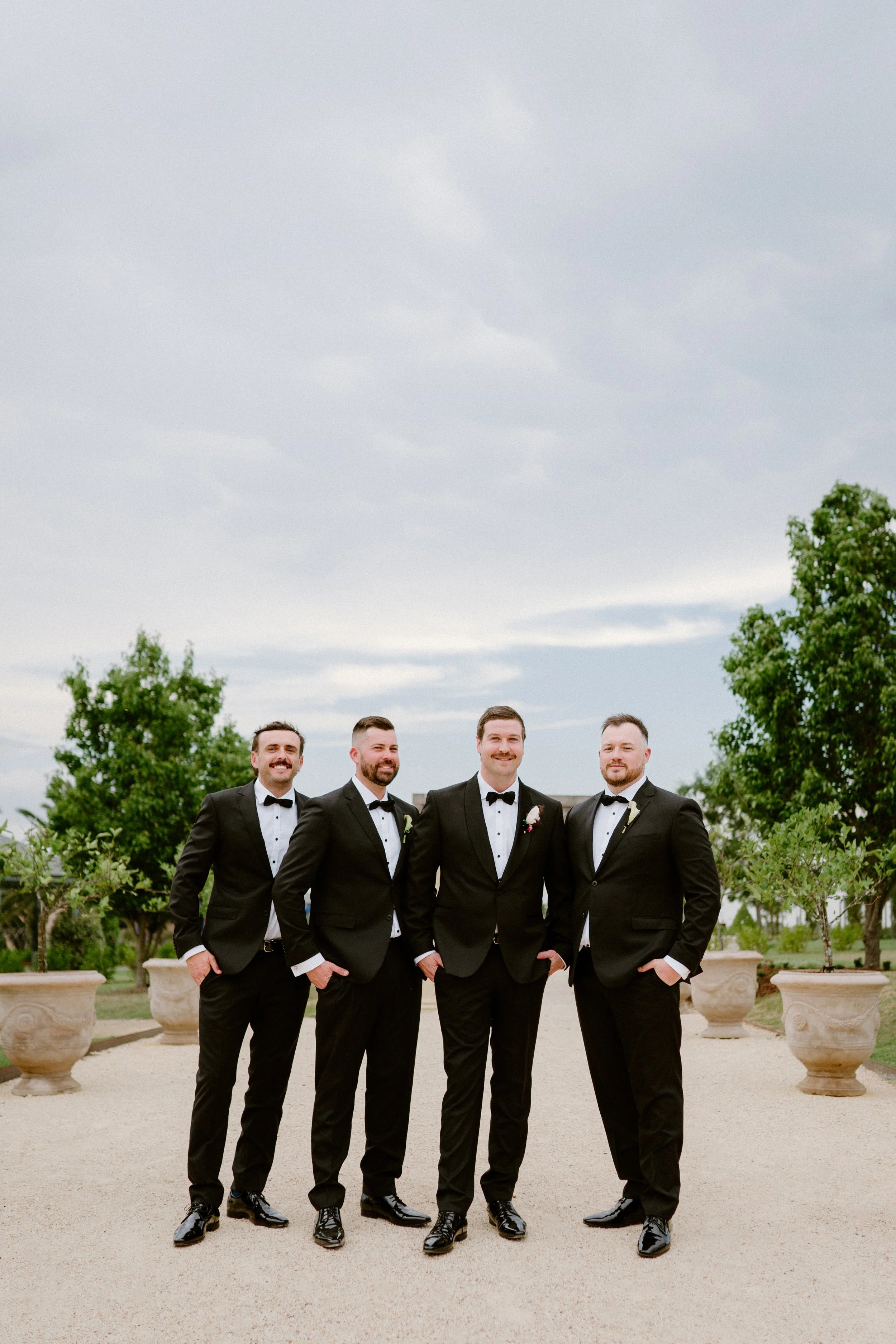 A group of four men in tuxedos standing outdoors on a gravel path, with trees and potted plants around them, and a cloudy sky overhead.