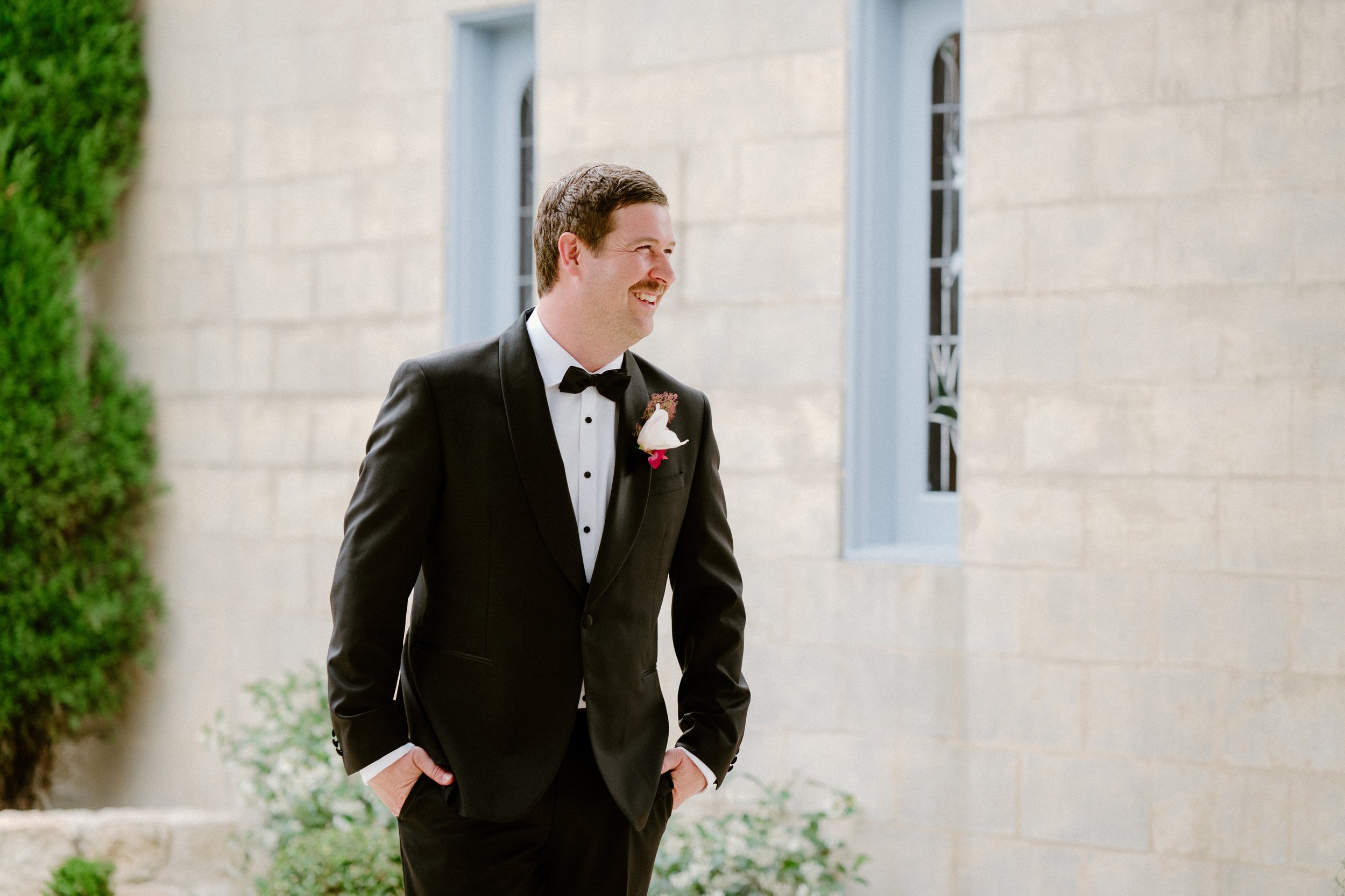A man dressed in a black tuxedo, black bow tie, and a boutonniere, smiling and looking to the side outside a building with light-colored stone walls and arched windows.