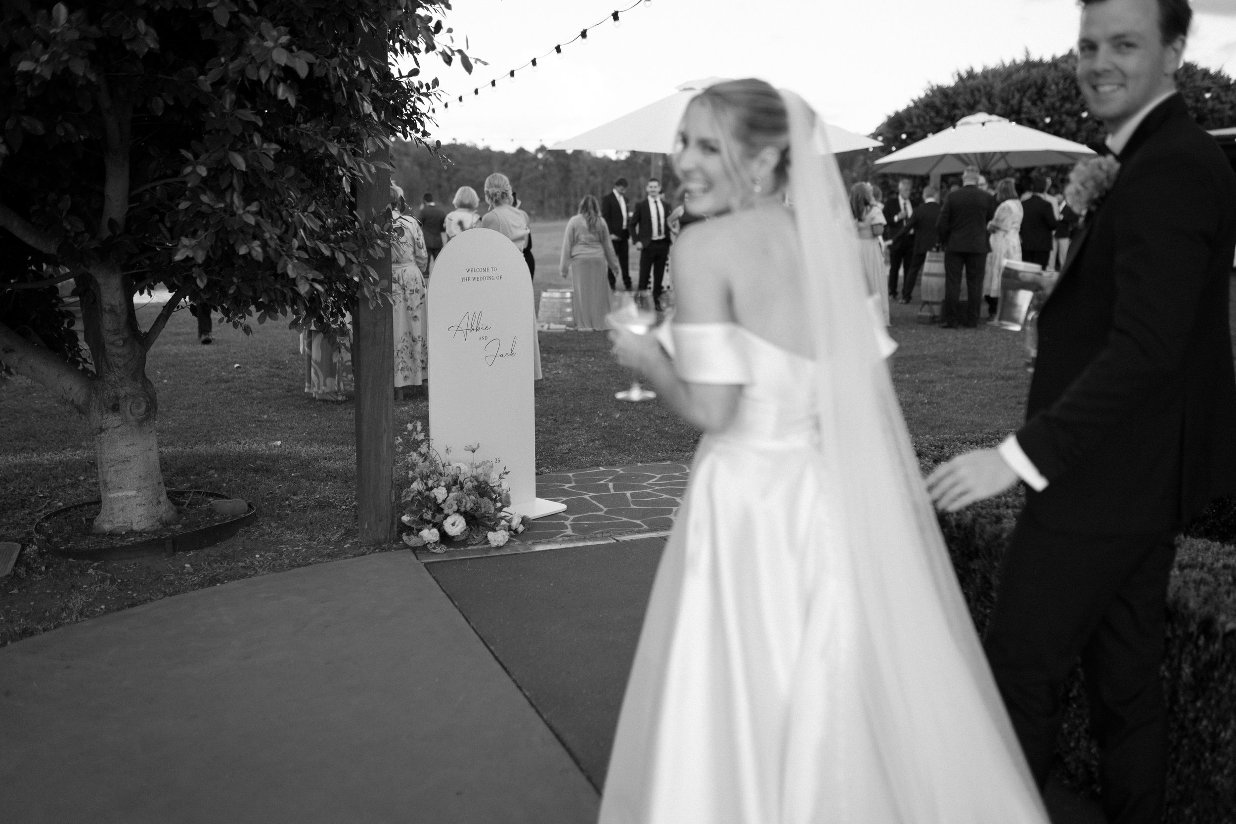 Black and white photo of a wedding reception outdoors with guests and outdoor umbrellas, a bride in a wedding dress holding a glass, a groom in a suit, and a wedding sign surrounded by flowers. Bimbadgen Palmers Lane Wedding, Hunter Valley