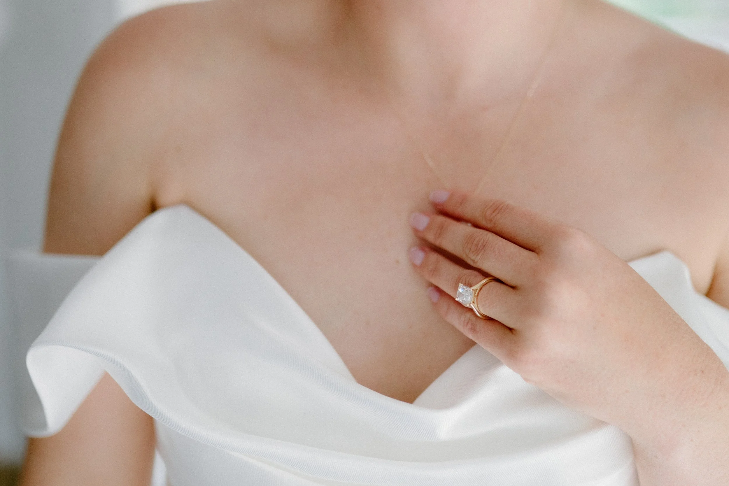 A woman wearing a white dress or top, with her hand on her chest showing a gold ring with a large, square diamond.