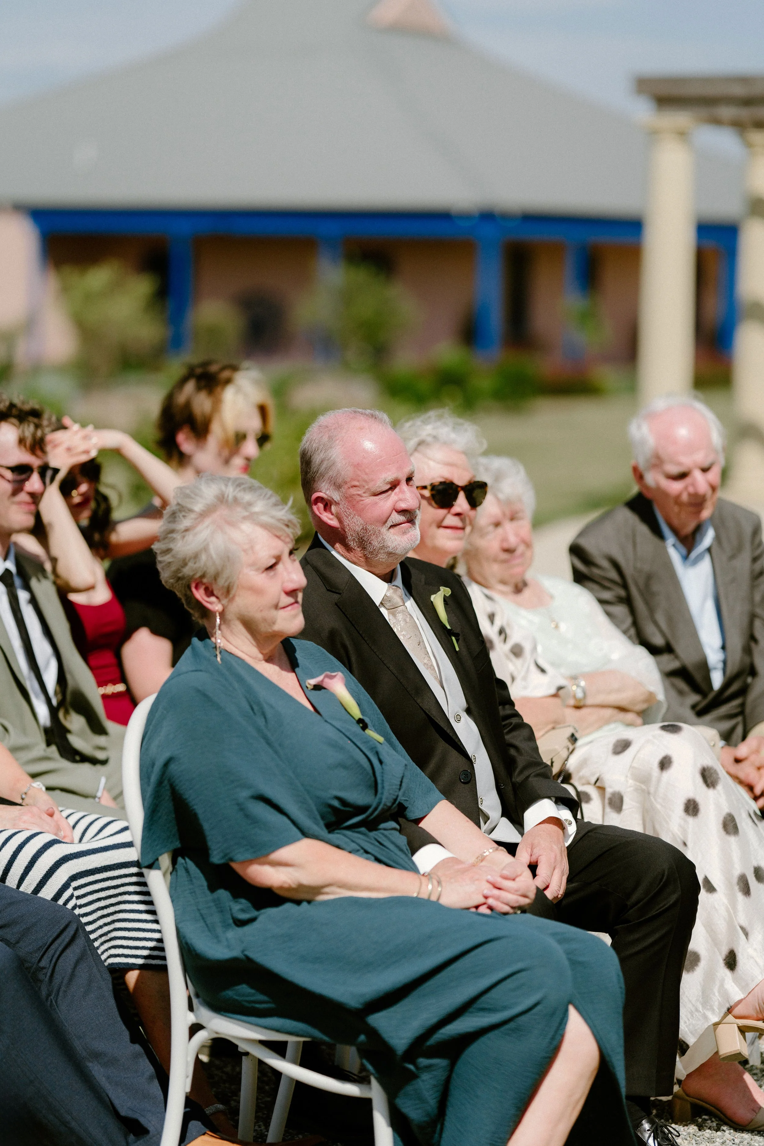 Group of elderly and young adults sitting outdoors at a formal event, likely a wedding, in sunlight, with a building and greenery in the background.
