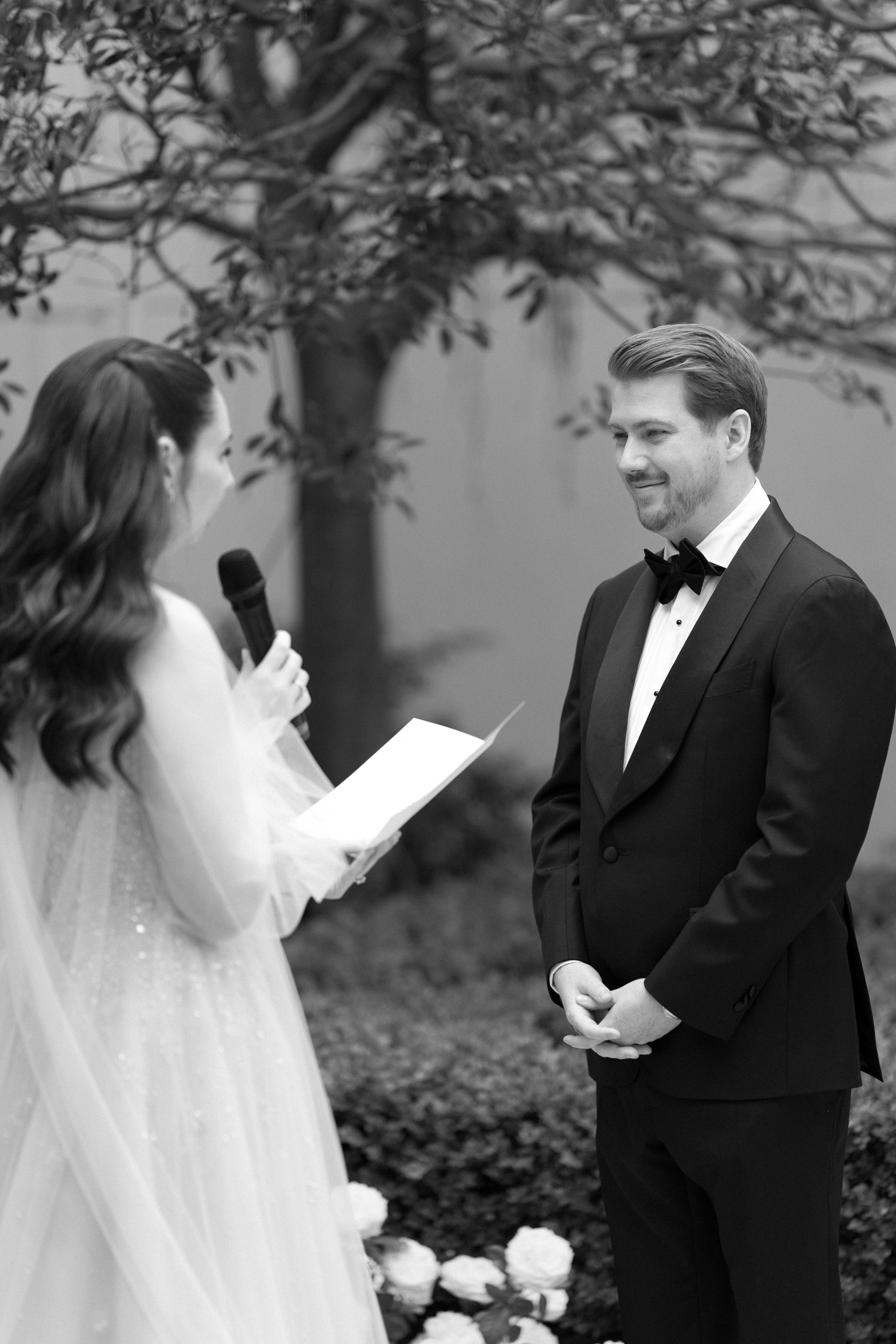 A woman in a wedding dress holding a microphone reads vows to a man in a tuxedo during a wedding ceremony outdoors, with a tree in the background.