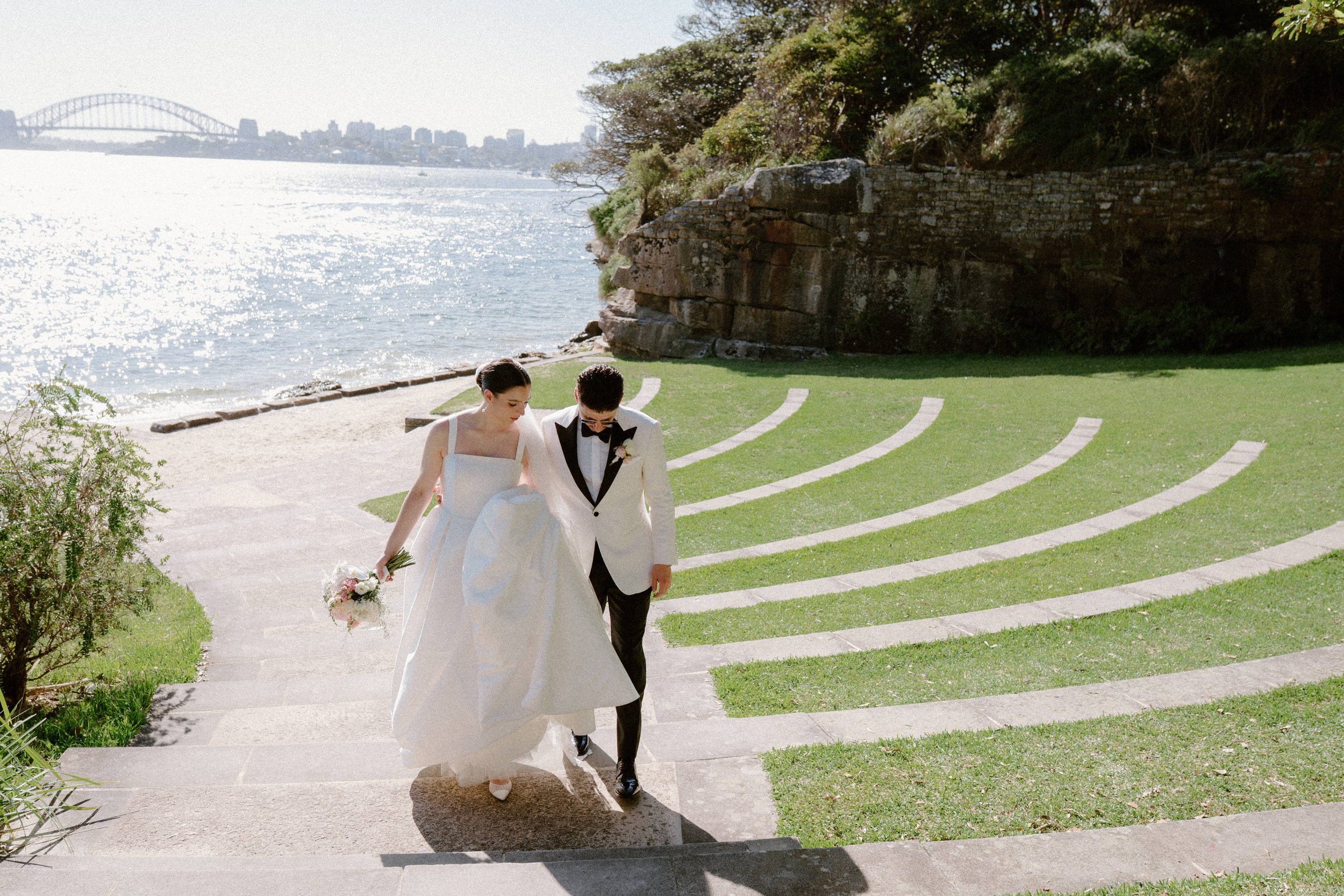 Bride and groom walking hand in hand down stone steps near a waterfront with a city and bridge in the background.