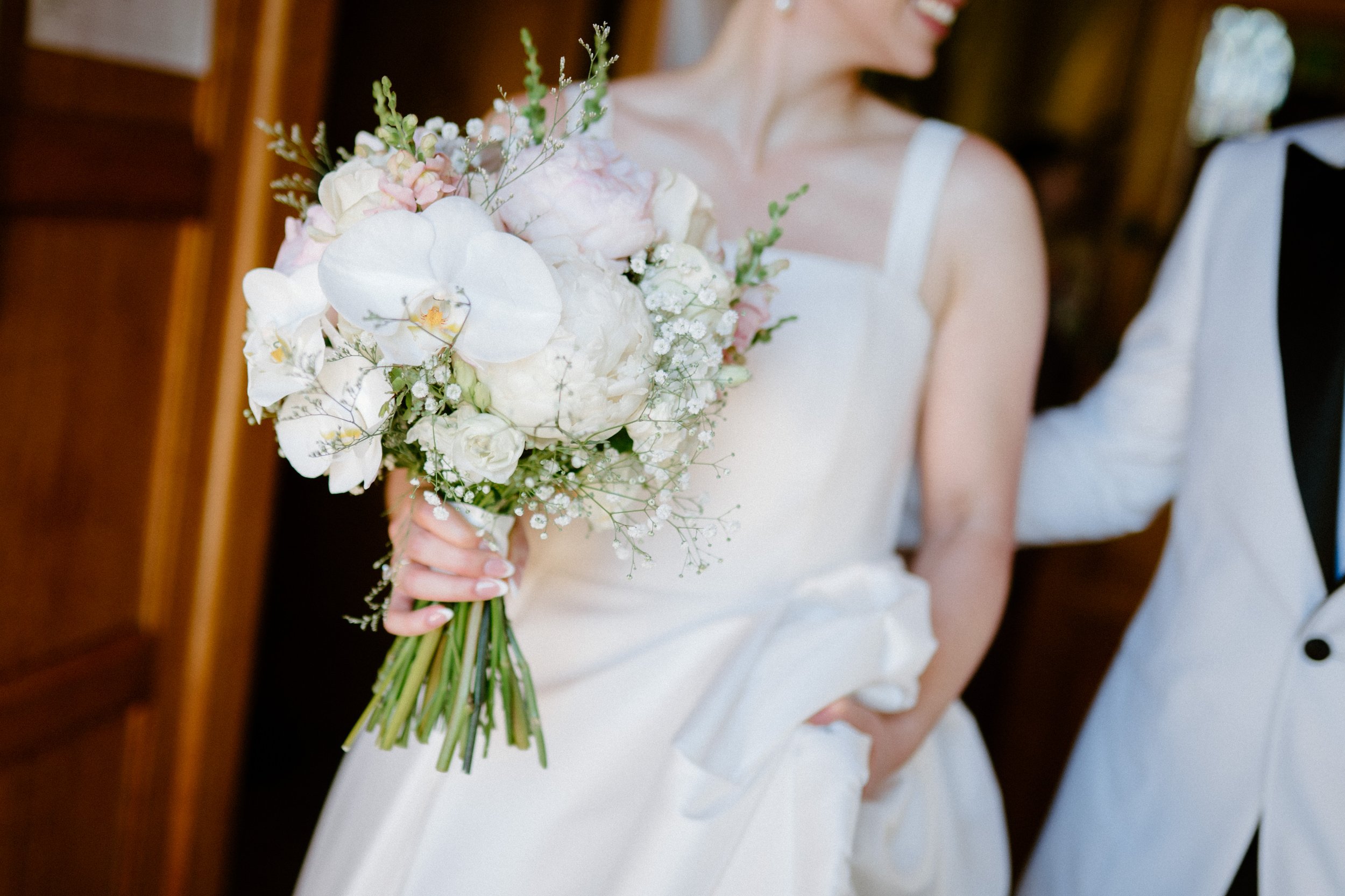 A bride holding a bouquet of white and pale pink flowers at her wedding. Part of her white dress and a man in a white tuxedo are visible.