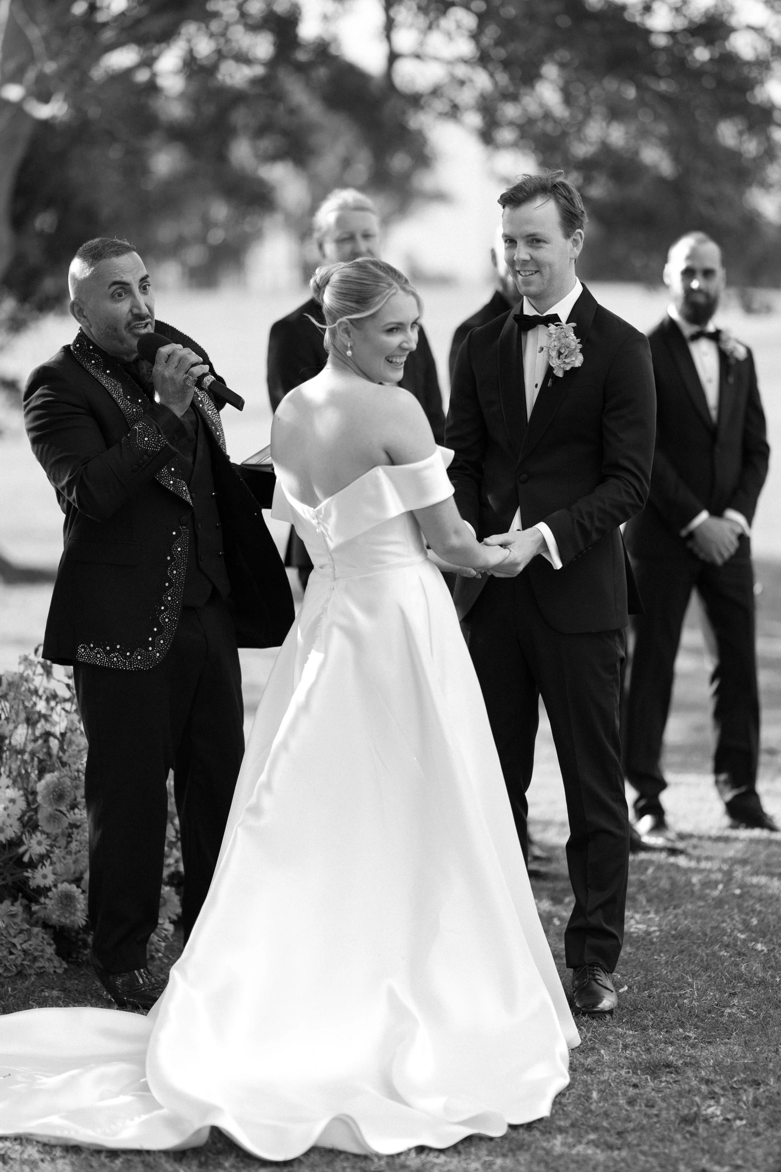 Wedding ceremony outdoors with bride and groom holding hands, officiant speaking, and groomsmen in background.