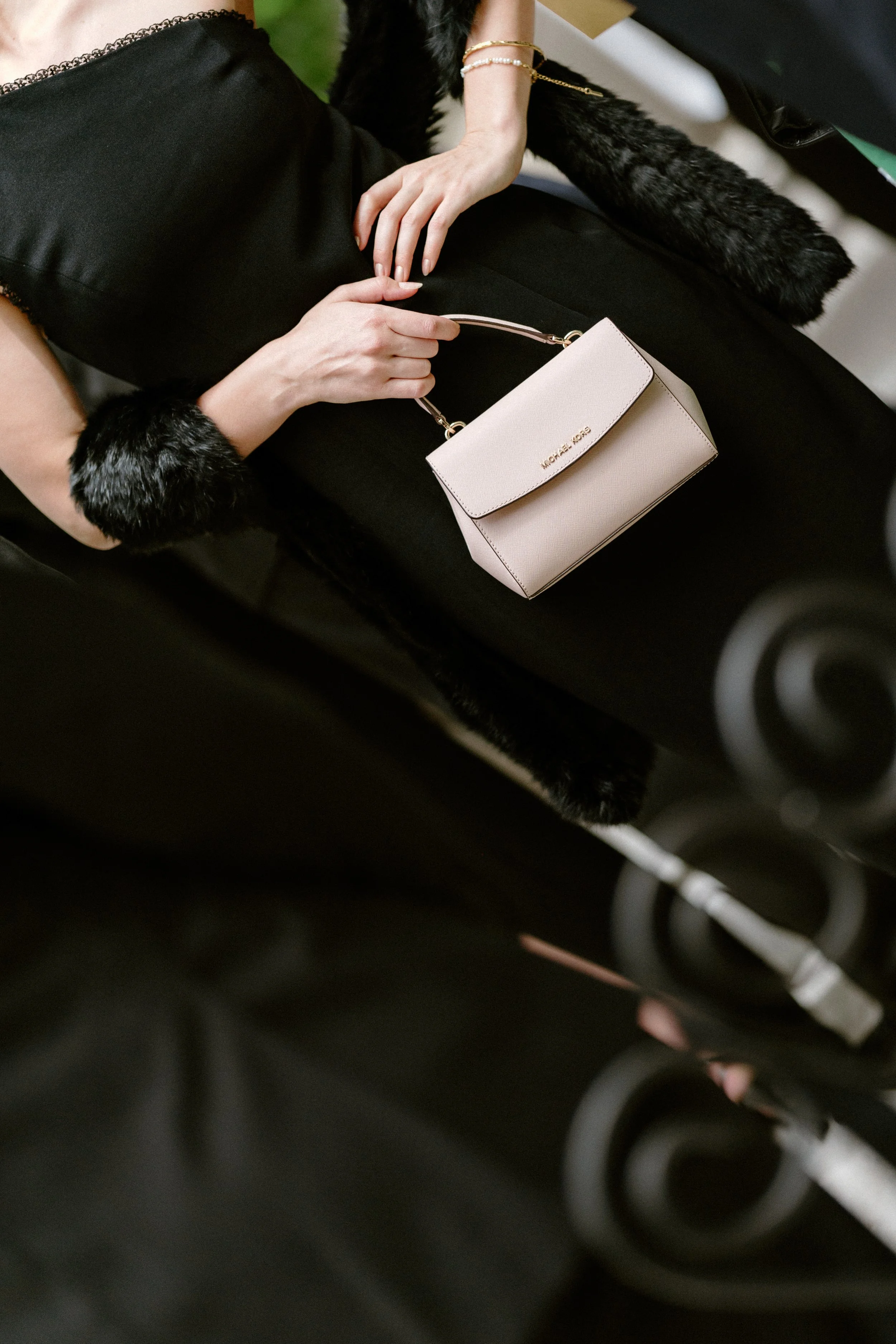 A woman dressed in black holding a light pink Micheal Kors handbag, seated in a black leather chair, with her hand resting on her lap, wearing pearl and gold bracelet accessories.