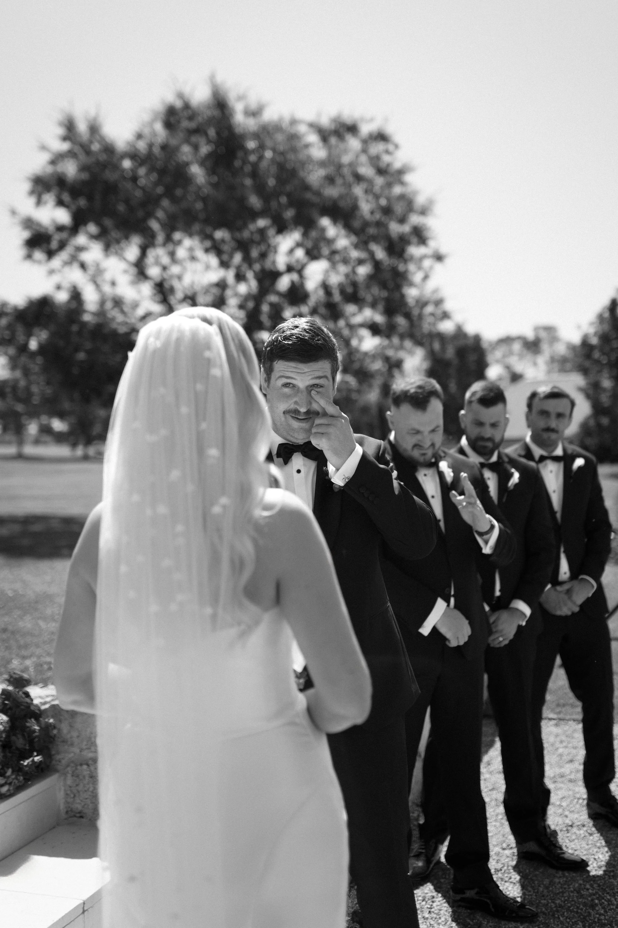 A groom in a tuxedo wiping his eye while talking to the bride, who is wearing a wedding gown and veil, during a wedding ceremony outdoors with groomsmen in suits smiling.