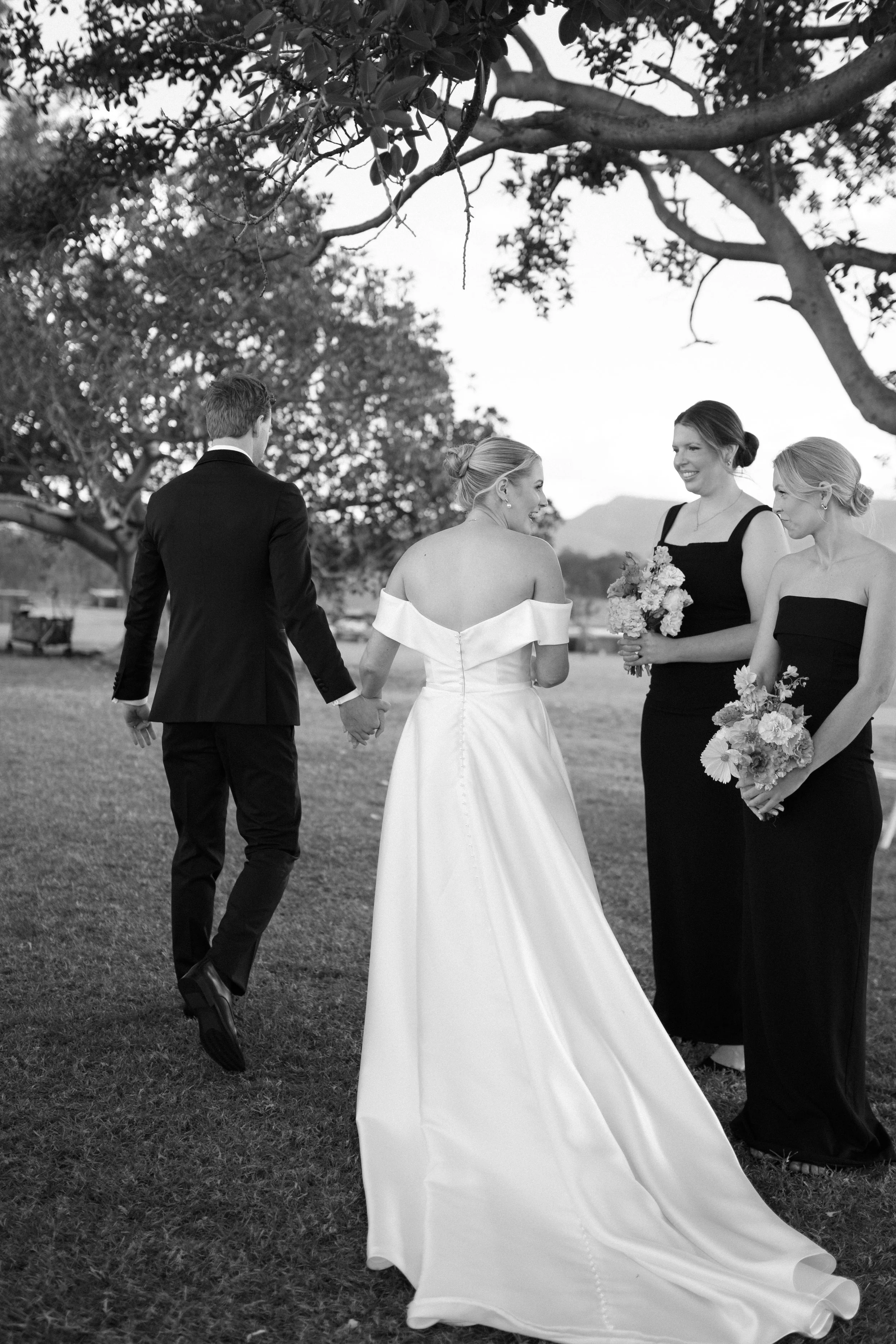 Black and white image of a wedding ceremony outdoors with three women and one man. The bride is wearing an off-shoulder wedding gown and is holding hands with the groom who is dressed in a dark suit. Two bridesmaids, dressed in black dresses, are sta