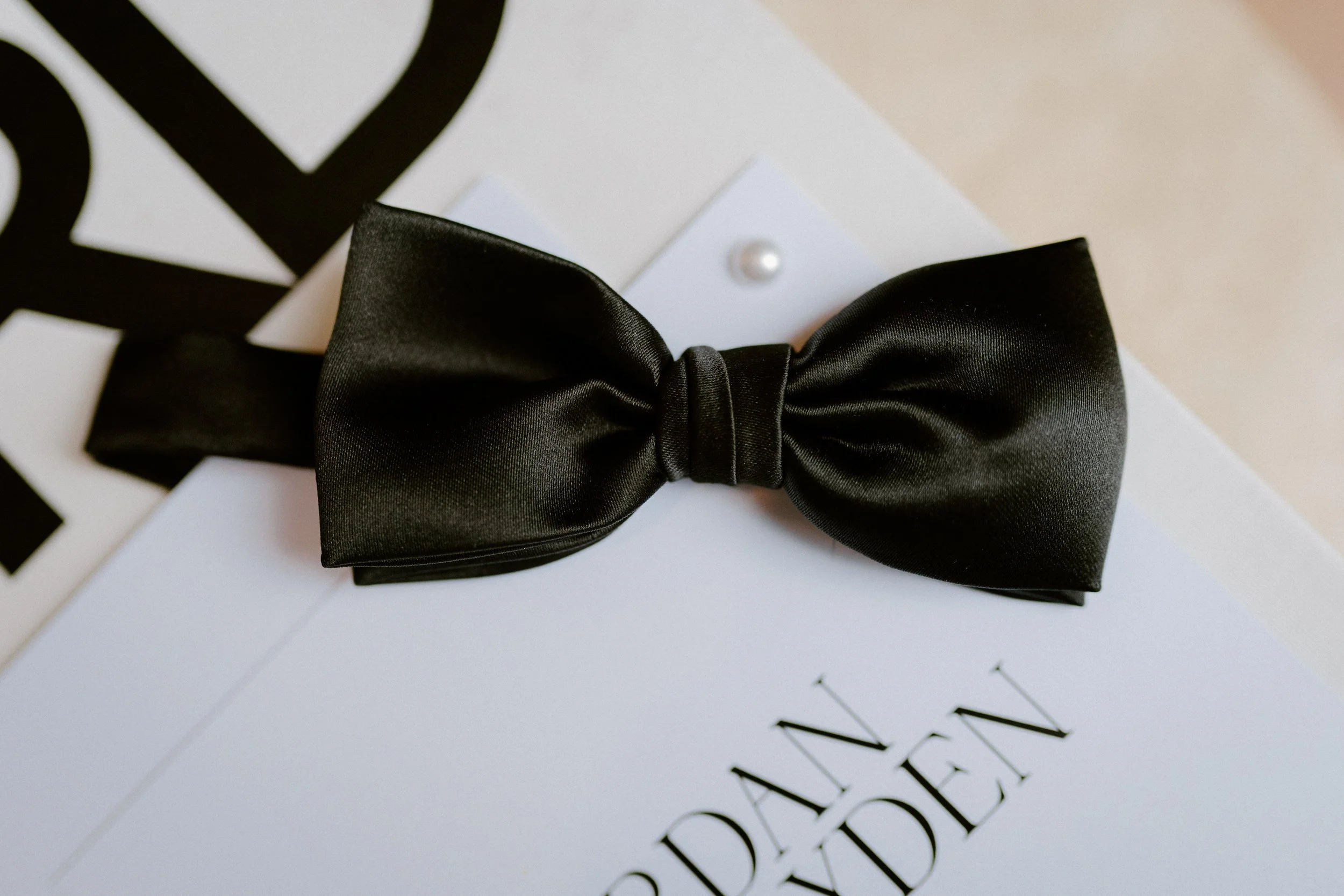 The Sienna, Hunter Valley Wedding Venue, Wedding Photos. Black satin bow tie resting on a white card with black text, pearl detail, and partially visible black paper with cut-out letters.