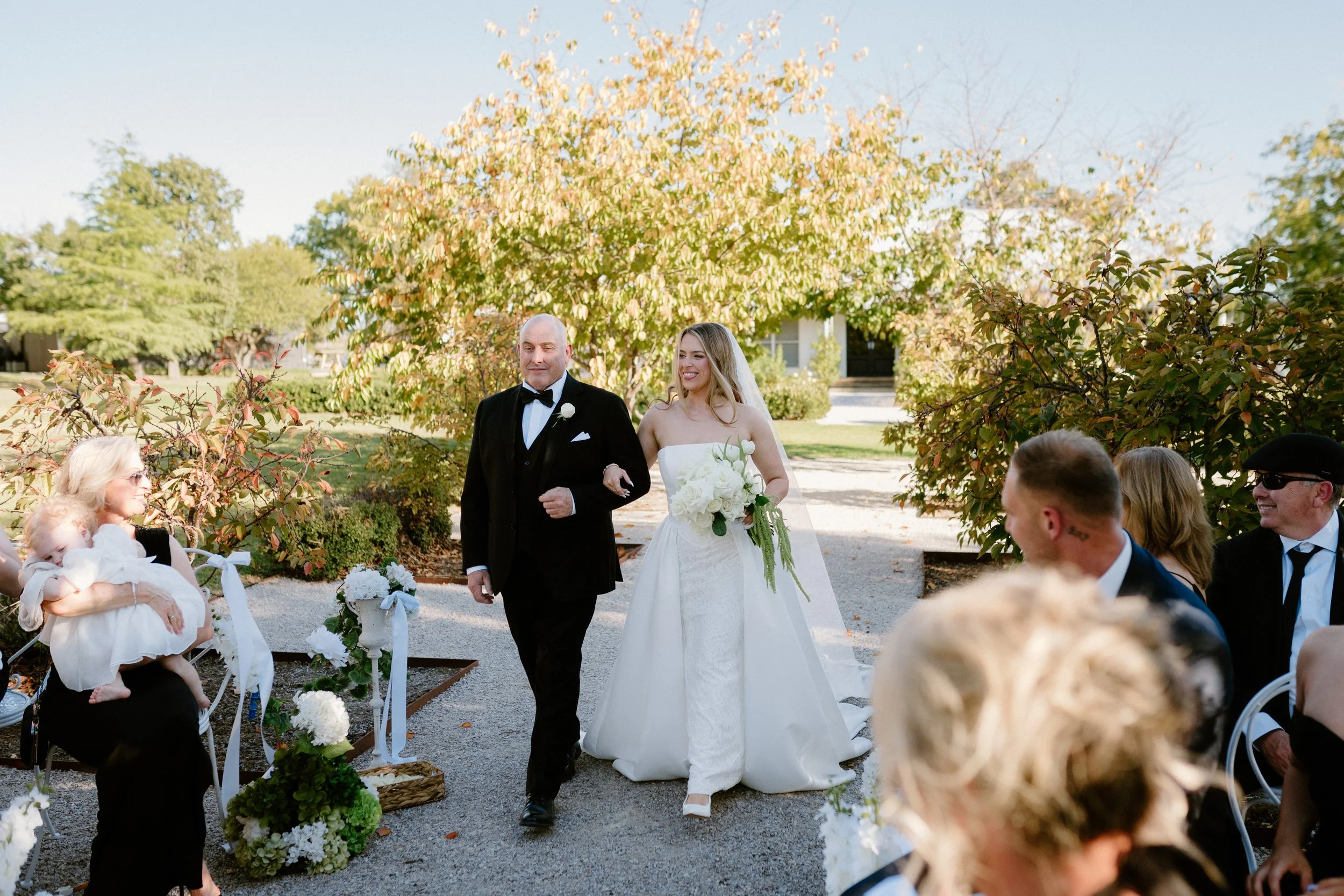 A bride in a white wedding dress and a groom in a black tuxedo walk down an outdoor aisle at a wedding ceremony, surrounded by seated guests and lush greenery.